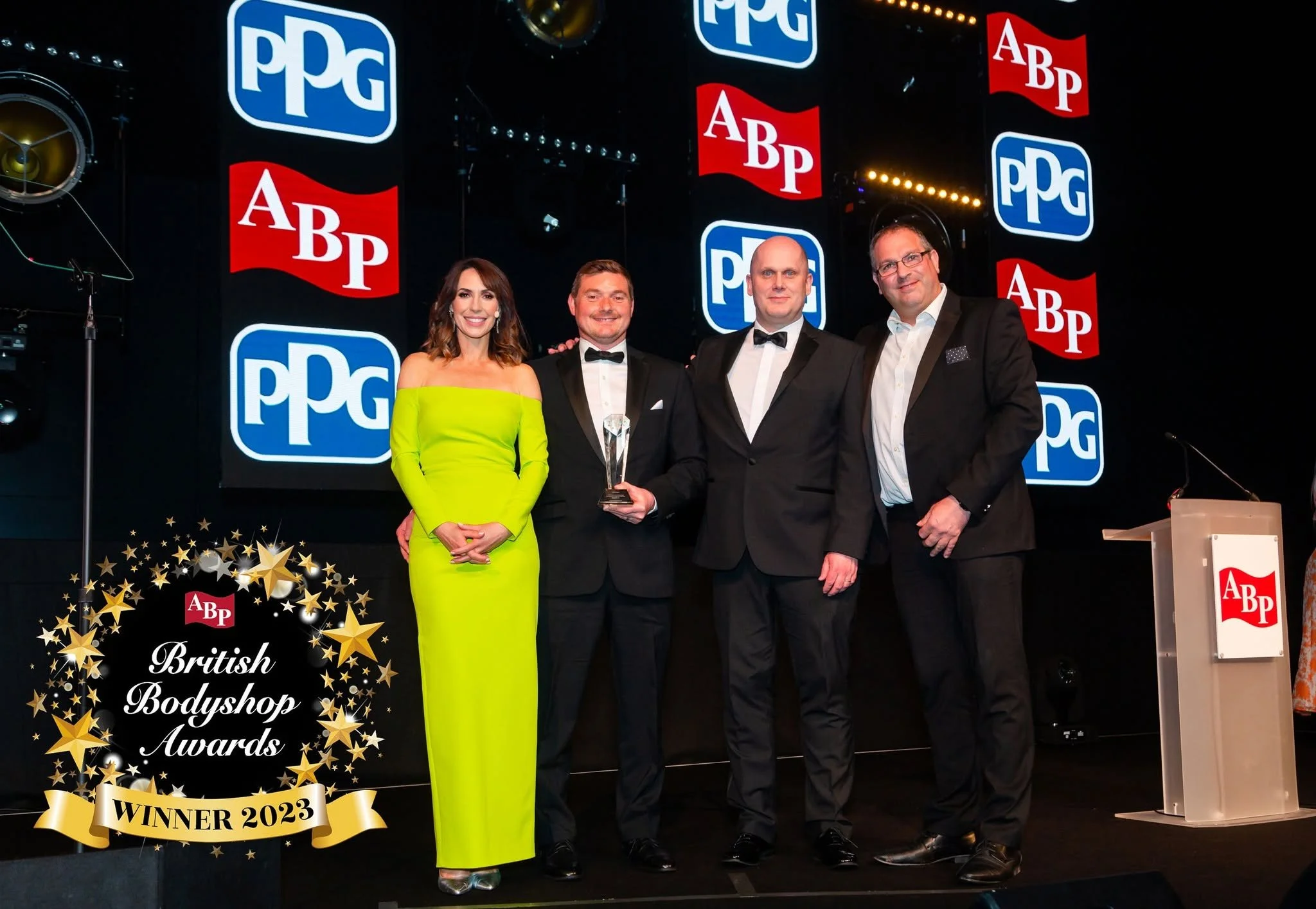 Four people in formal attire posing on stage, holding an award, at the British Bodyshop Awards. The background displays multiple logos for PPG and ABP. One woman is wearing a bright yellow off-the-shoulder gown, and three men are dressed in tuxedos. A banner in the foreground reads 'Winner 2023' with stars.