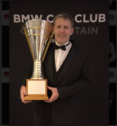 A man in a tuxedo holding a large trophy at a BMW Club event.