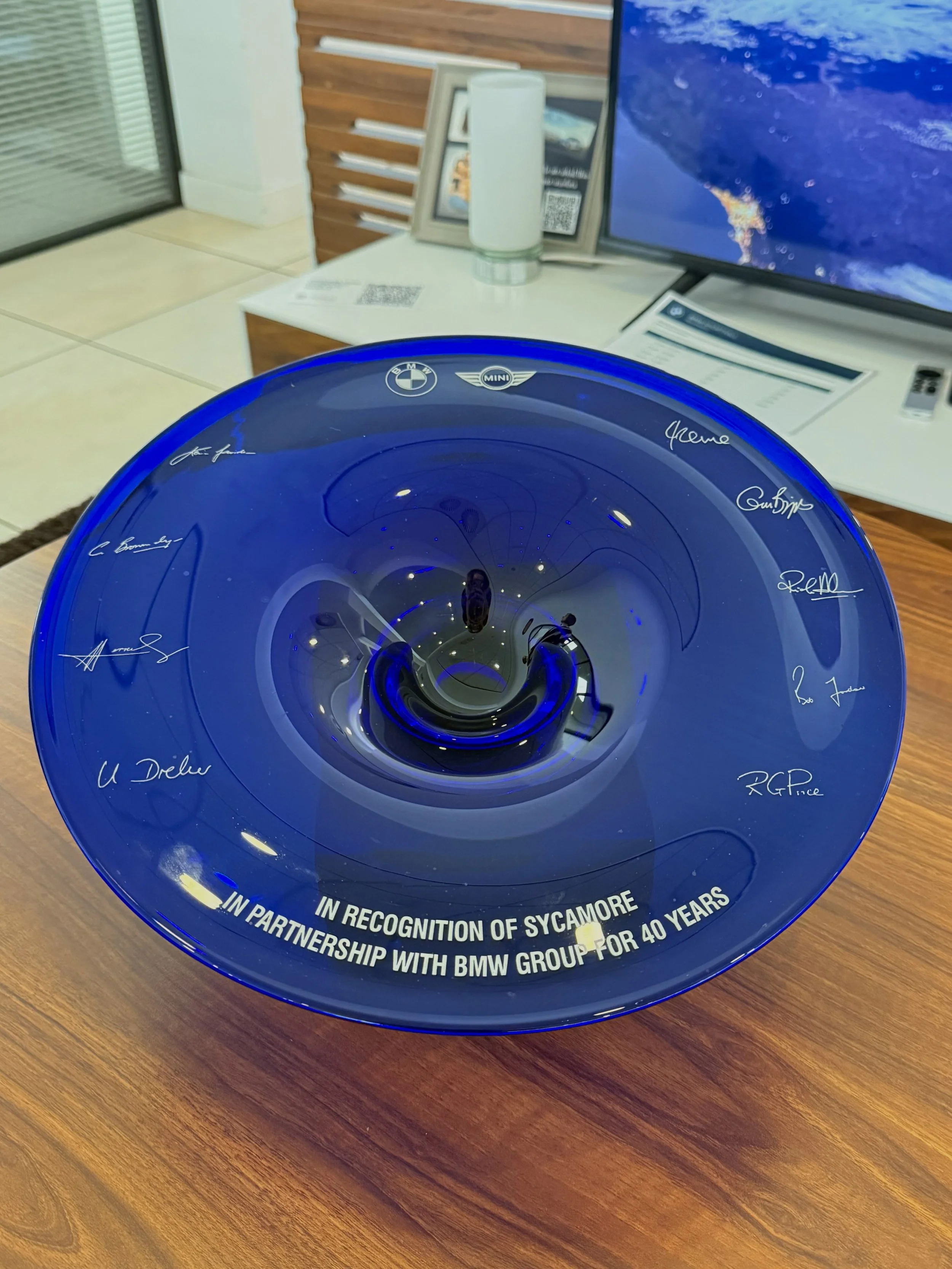 Blue circular award plaque with signatures, BMW and MINI logos, and white text that reads: "IN PARTNERSHIP WITH BMW GROUP FOR 40 YEARS IN RECOGNITION OF SYCAMORE" placed on a wooden surface, with an office background including a computer monitor, paperwork, and other office items.