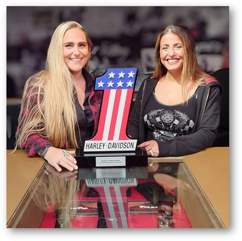 Two women smiling and holding a Harley-Davidson sign with an American flag design. They are seated at a glass display case with reflections visible.