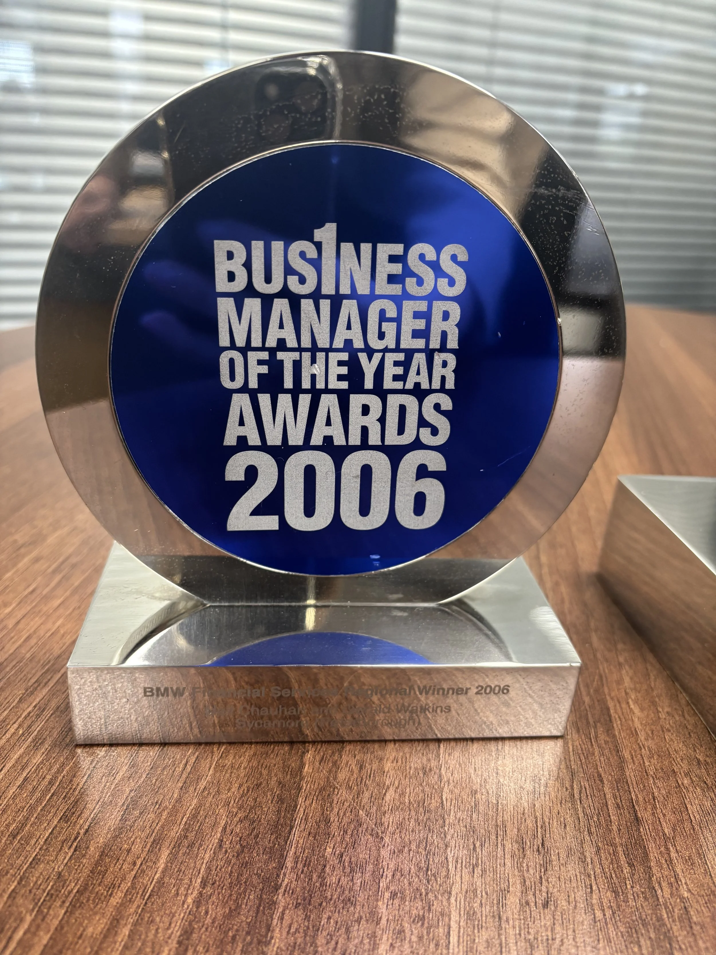A shiny silver award with a blue circular sticker in the center reading 'Business Manager of the Year Awards 2006'. It is placed on a wooden surface with a background of window blinds.
