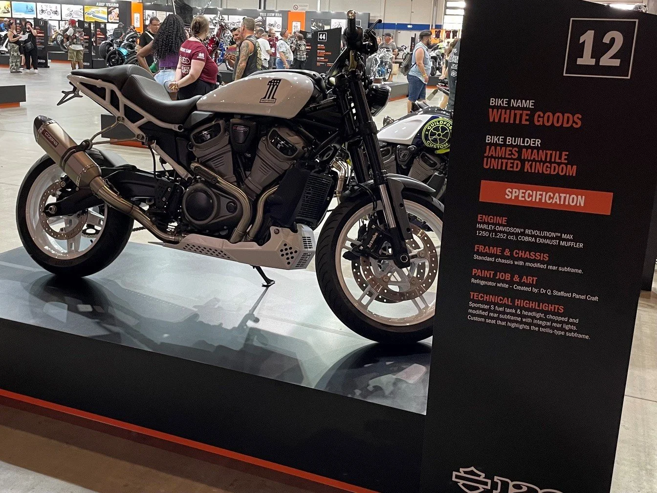A white custom Harley-Davidson motorcycle displayed at a motorcycle exhibition with a black background sign providing specifications, including engine type, frame and chassis details, paint job, and technical highlights.