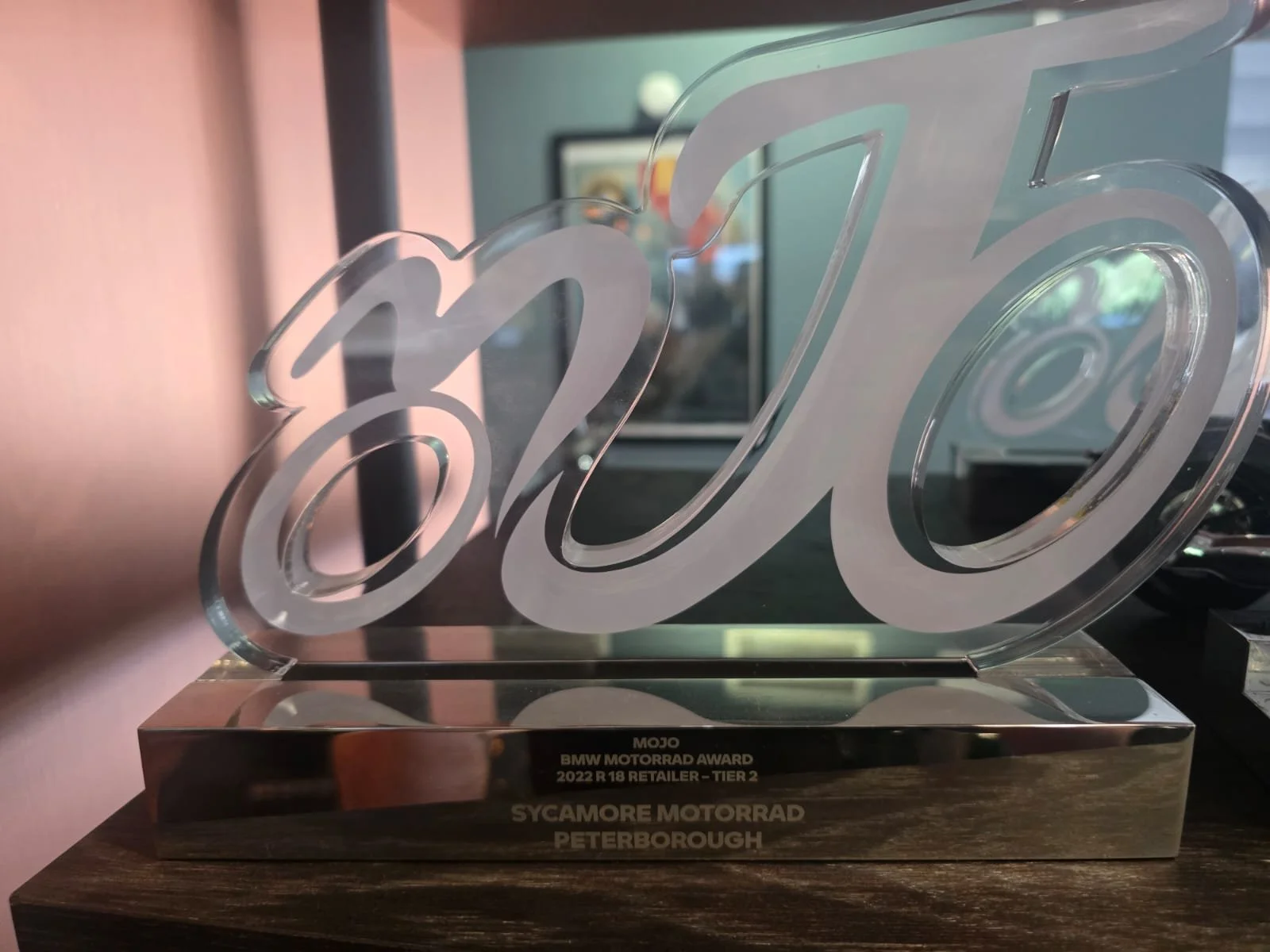A trophy with the BMW logo, engraved with 'MOJO BMW MOTORRAD AWARD 2022 R 18 RETAILER - TIER 2' and 'SYCAMORE MOTORRAD PETERBOROUGH', displayed on a dark wooden surface.