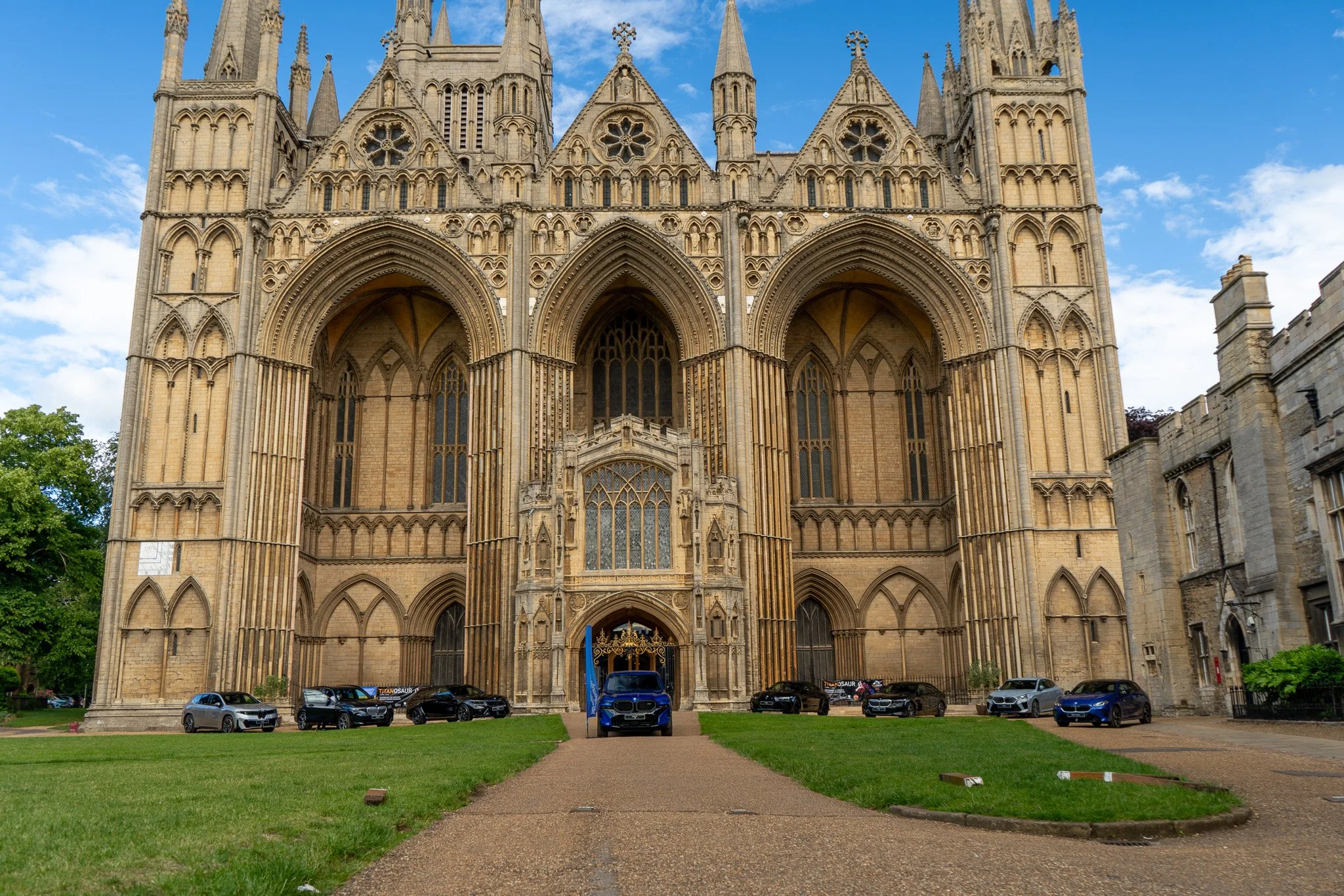 A large, historic Gothic cathedral with elaborate stone architecture, pointed arches, and detailed windows, situated behind a grassy area with parked cars and a paved walkway leading up to its entrance, under a blue sky with scattered clouds.