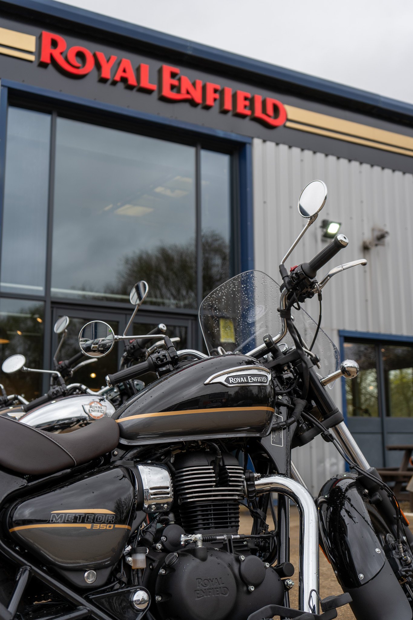 Close-up view of several black Royal Enfield motorcycles parked outside a building with a sign that reads 'Royal Enfield,' featuring a red logo and blue frame, under a cloudy sky.