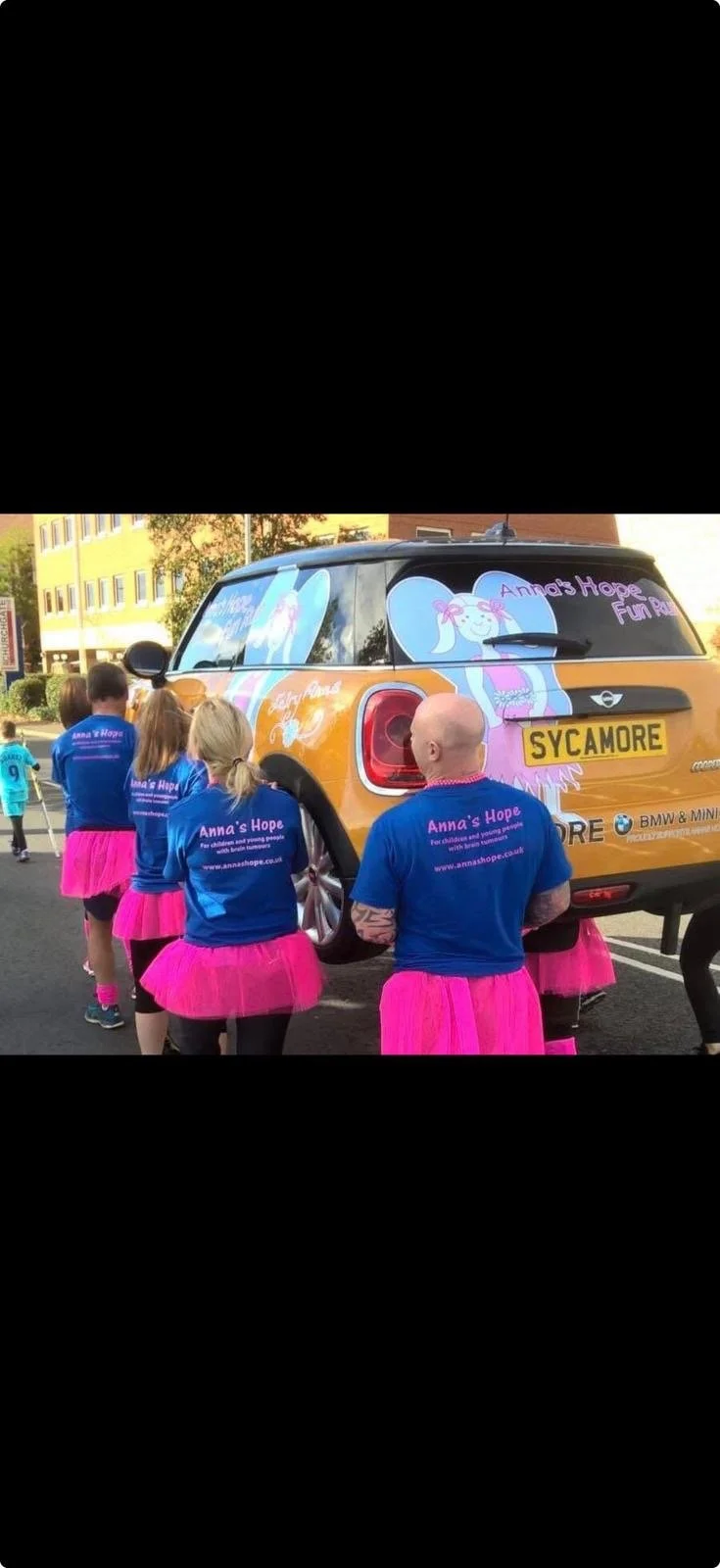People wearing blue shirts and pink tutus surrounding a Mini Cooper car with Anna's Hope charity branding.