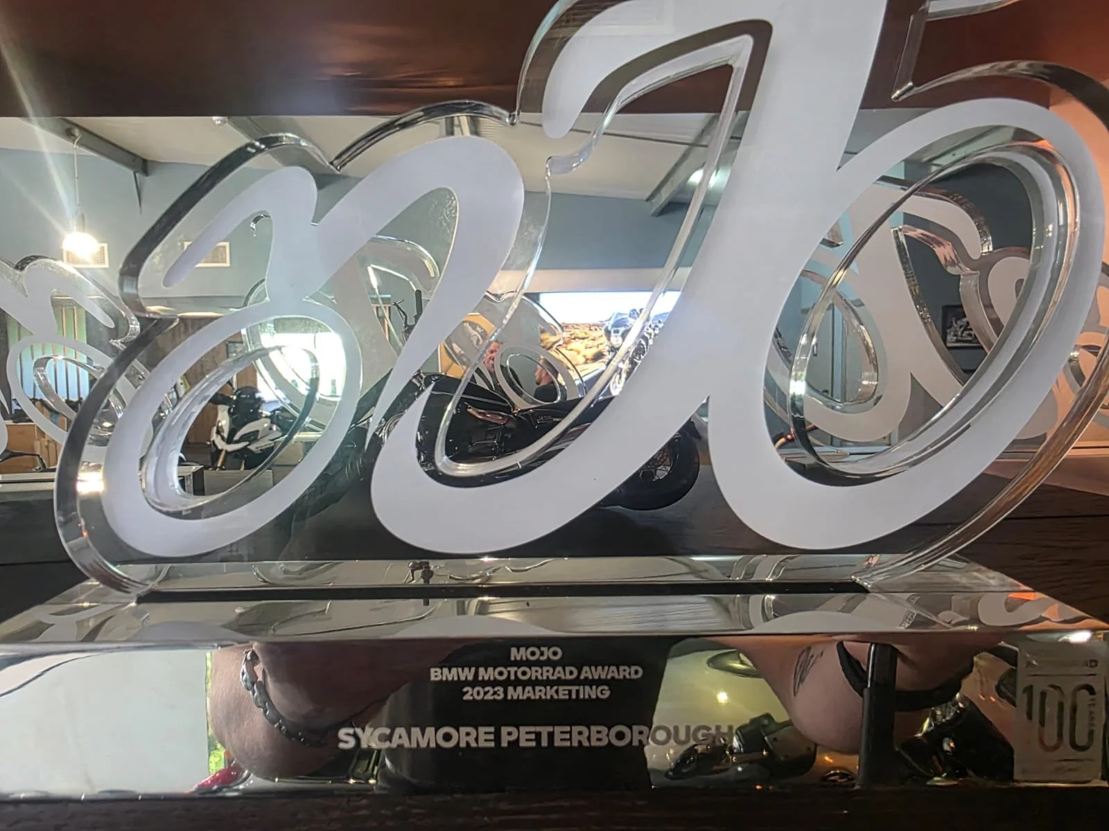 A clear acrylic award plaque with stylized cursive lettering and silver accents, featuring inscriptions for the 2023 BMW Motorrad Award, presented at a marketing event in Sycamore, Peterborough.