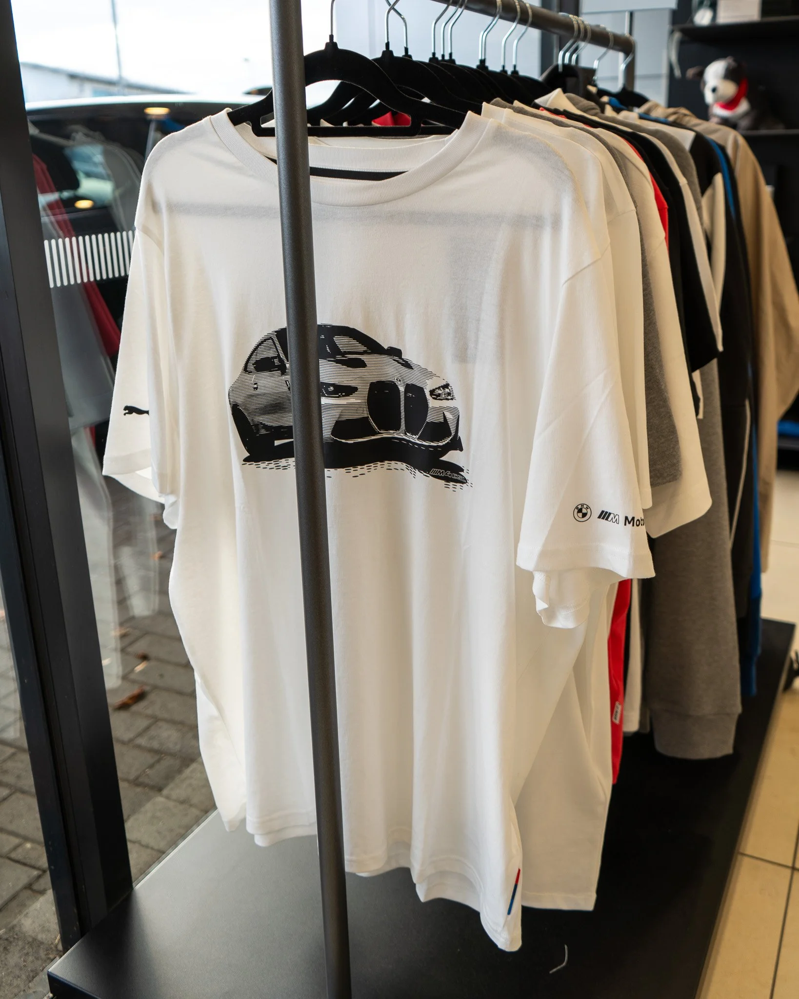 White T-shirt with a black graphic of a sports car on the front, hanging on a black hanger in a clothing store. Other shirts and clothing items are visible in the background.