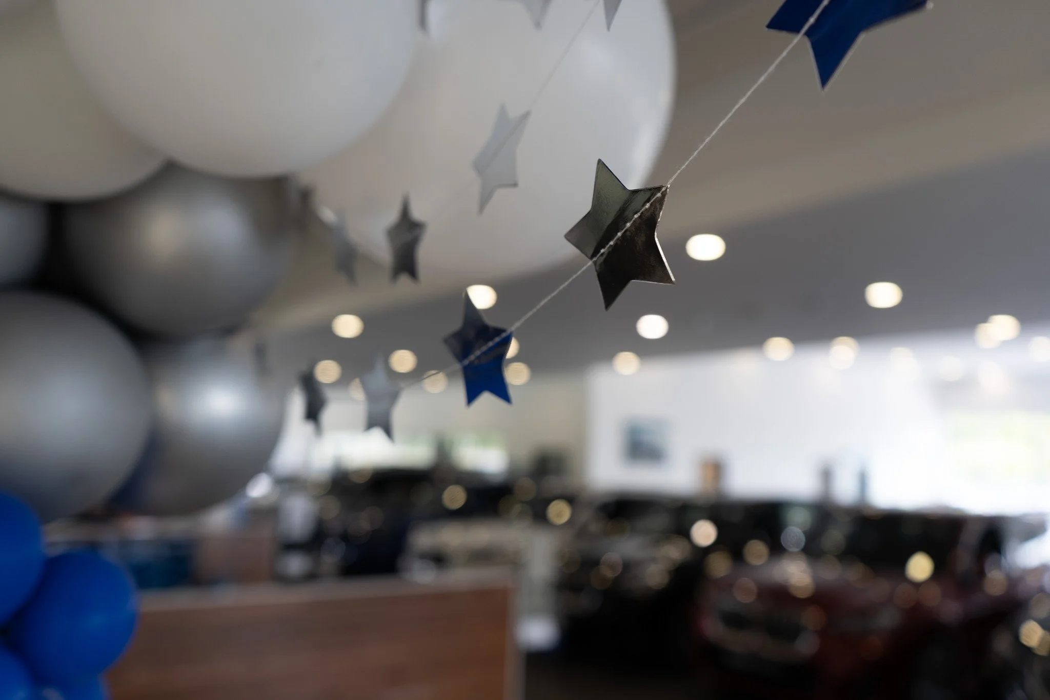 White and silver balloons with star-shaped hanging decorations, some blue balloons visible, blurred background with circular lights.