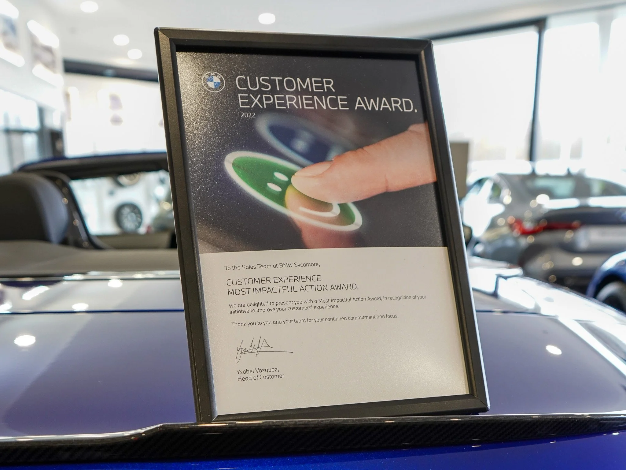 A framed certificate awarded for the Most Impactful Action, placed on a car's dashboard with a blurred car showroom in the background.