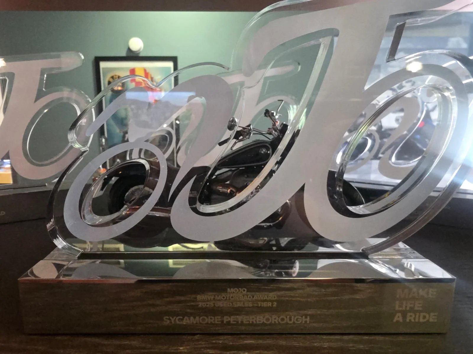 A transparent award with artistic lettering, featuring a small motorcycle inside, on a wooden base with engraved text.