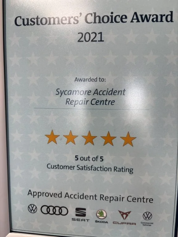 Photograph of a service award certificate from 2021 given to Sycamore Accident Repair Centre, showing a 5 out of 5 customer satisfaction rating with five gold stars, and logos of Volkswagen, Audi, Seat, Skoda, Cupra, and Volkswagen Commercial Vehicles at the bottom.