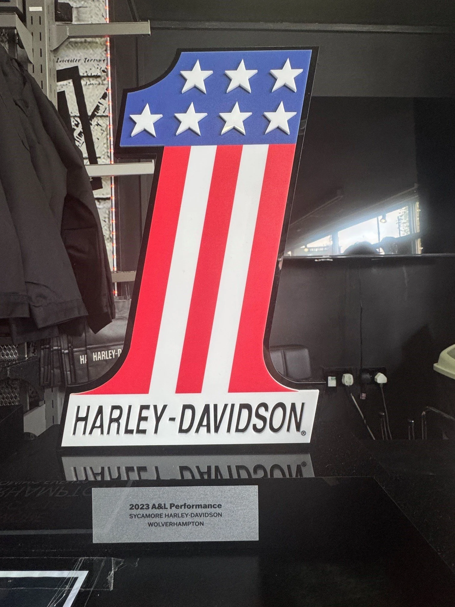 Harley-Davidson motorcycle dealership sign with red, white, and blue colors, featuring a large number 1 with five stars on top and the Harley-Davidson logo at the bottom. A smaller sign below reads "2023 A&L Performance, Sycamore Harley-Davidson, Wolverhampton."