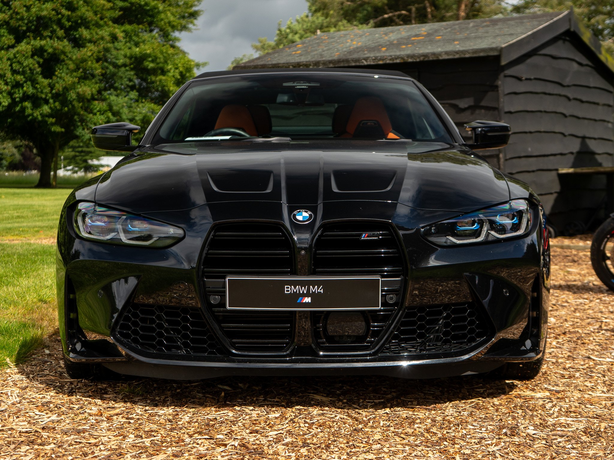 Front view of a black BMW M4 parked outdoors on a grassy area with trees and a rustic shed in the background.