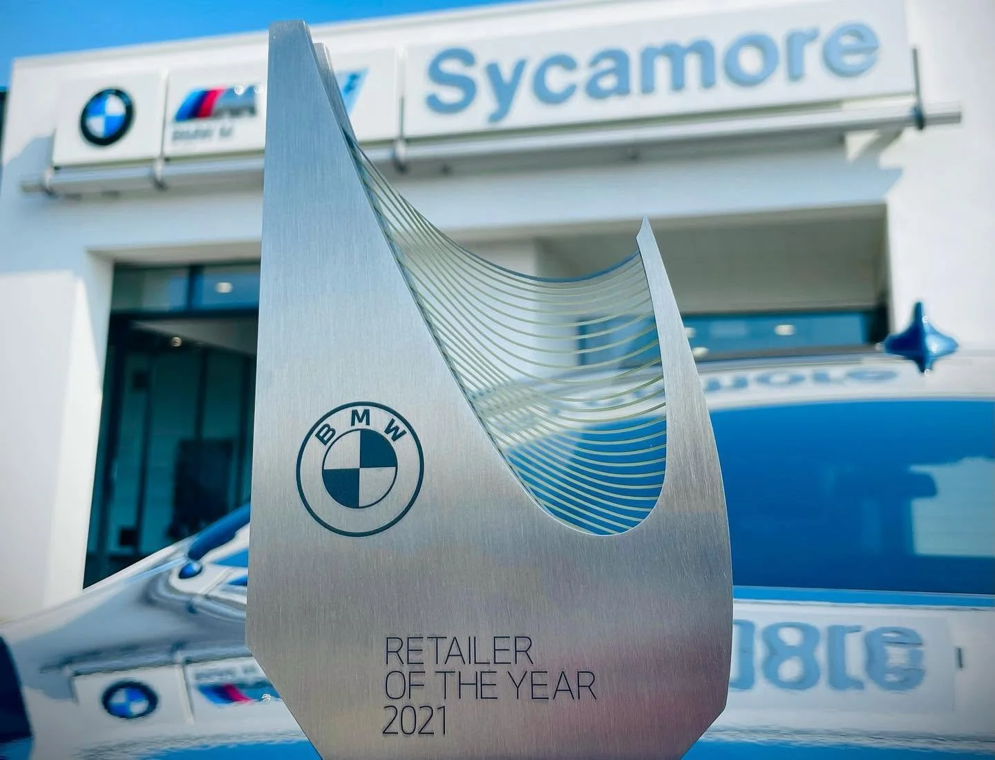 A silver BMW award titled "Retiler of the Year 2021" in front of a BMW dealership with Sycamore signage and BMW, BMW M, and BMW i logos.
