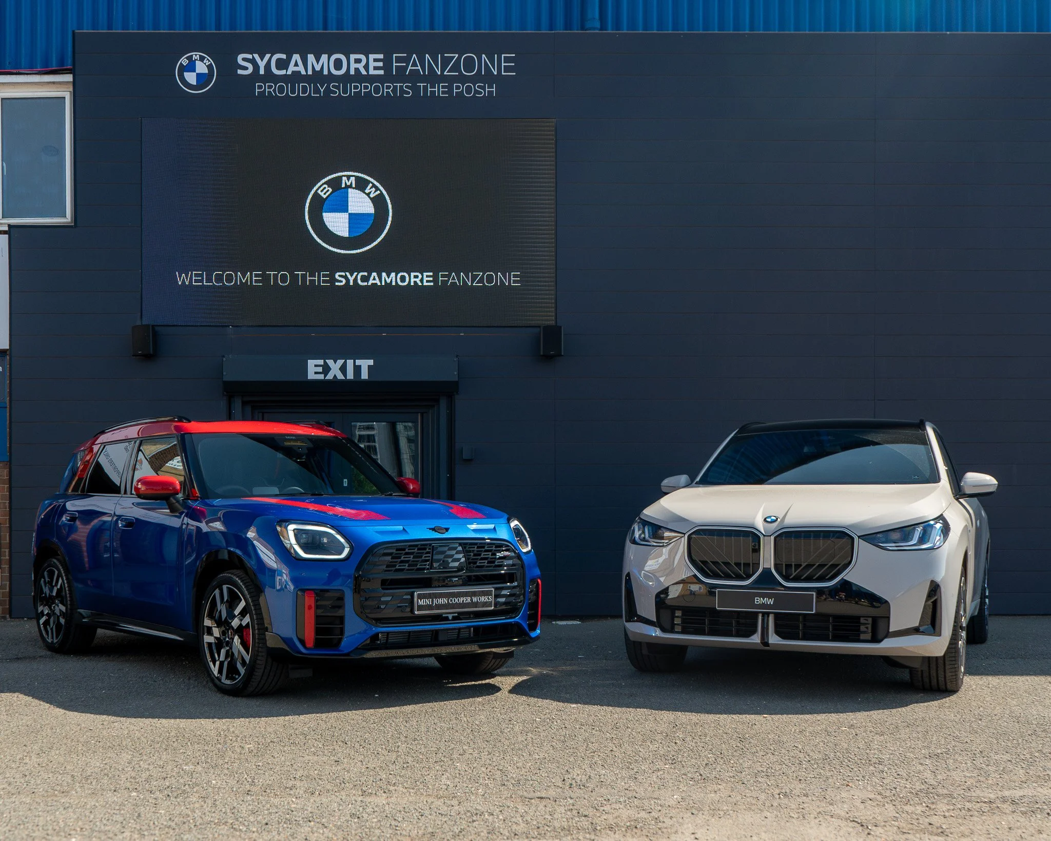 A car dealership parking lot with three cars in front of a building. The building has signs for 'Sycamore Fanzone,' BMW, Mini John Cooper Works, and BMW. There is an exit sign above a door. The sky is clear and the lighting suggests it is daytime.