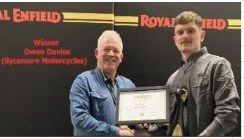 Two men standing on a stage, one holding a framed certificate, with a backdrop displaying the Royal Enfield and winner Owen Davies from Sycamore Motorcycles.