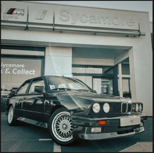 Black BMW M3 parked outside Sycamore dealership, close-up shot showing front and side view, with Sycamore signage above the entrance.