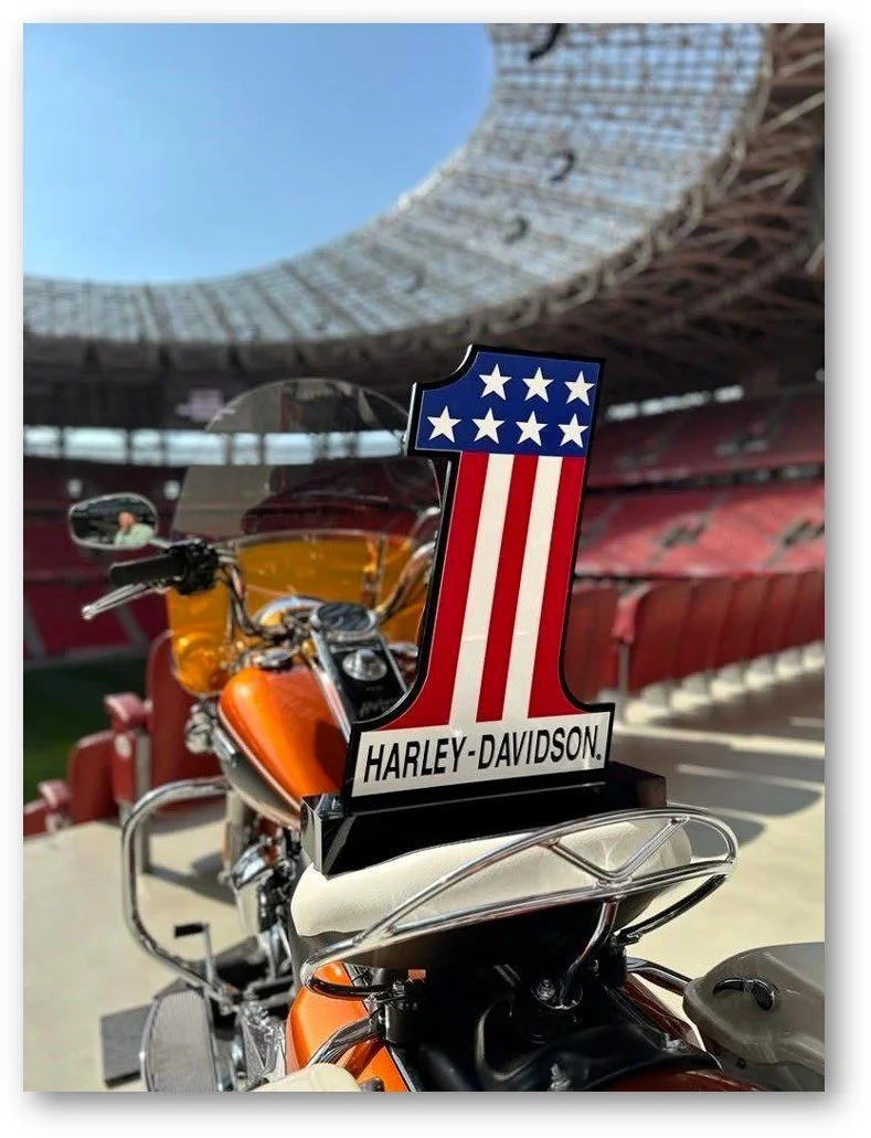 Harley-Davidson motorcycle with a patriotic American flag-themed accessory on a stadium motorcycle ramp, with an empty red seating area and an open roof opening in the background.
