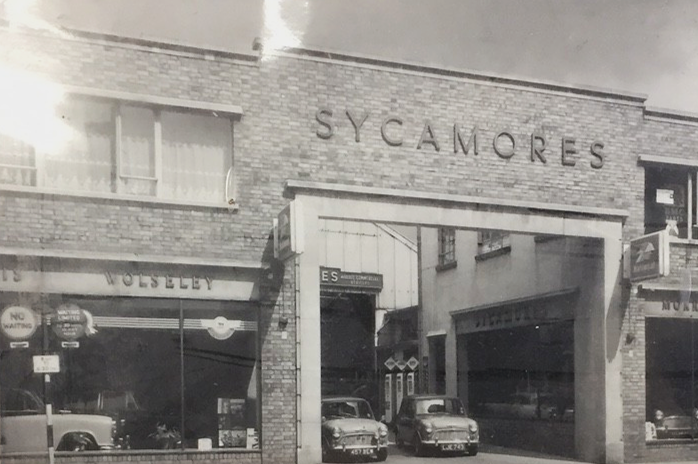 The front of a building with the word 'SYCAMORES' written on it, showing an entrance with two vintage cars parked in front.