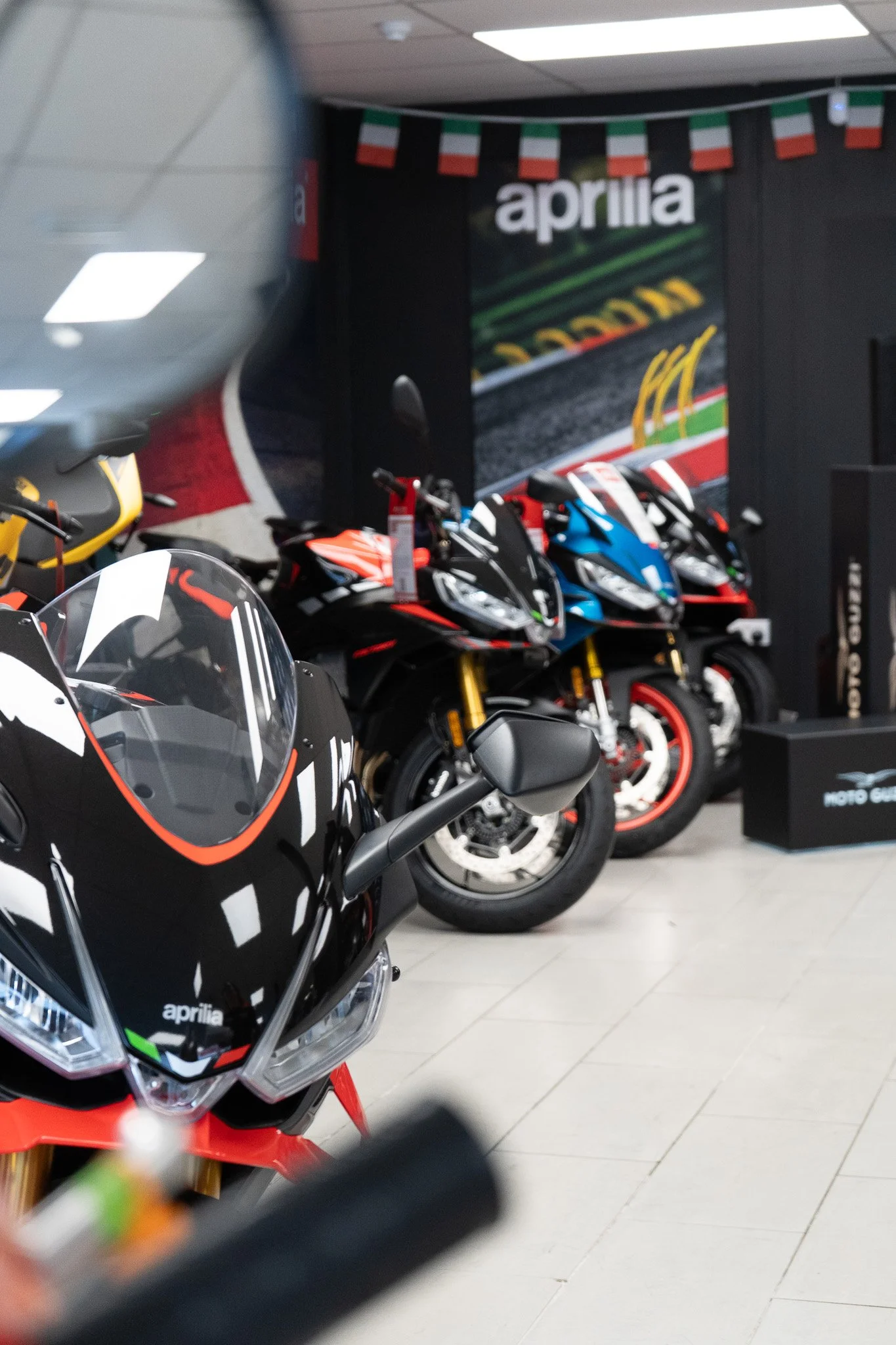 A row of Aprilia motorcycles on display inside a showroom with Italian flags hanging from the ceiling.