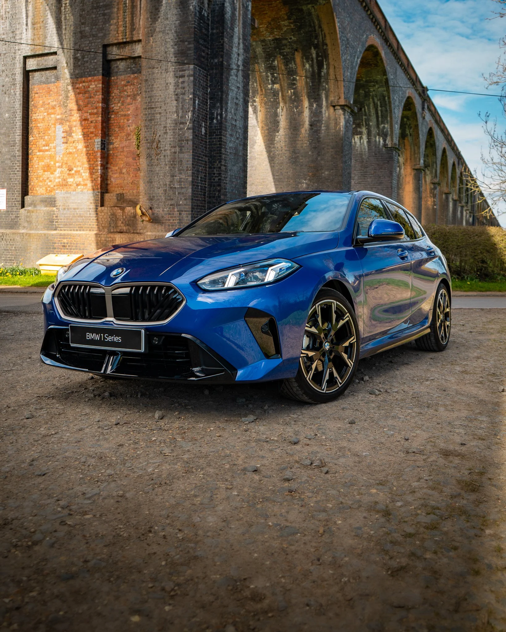A blue BMW 1 Series car parked on a dirt surface in front of an old brick aqueduct with multiple arches. The sky is partly cloudy with some visible trees and greenery.