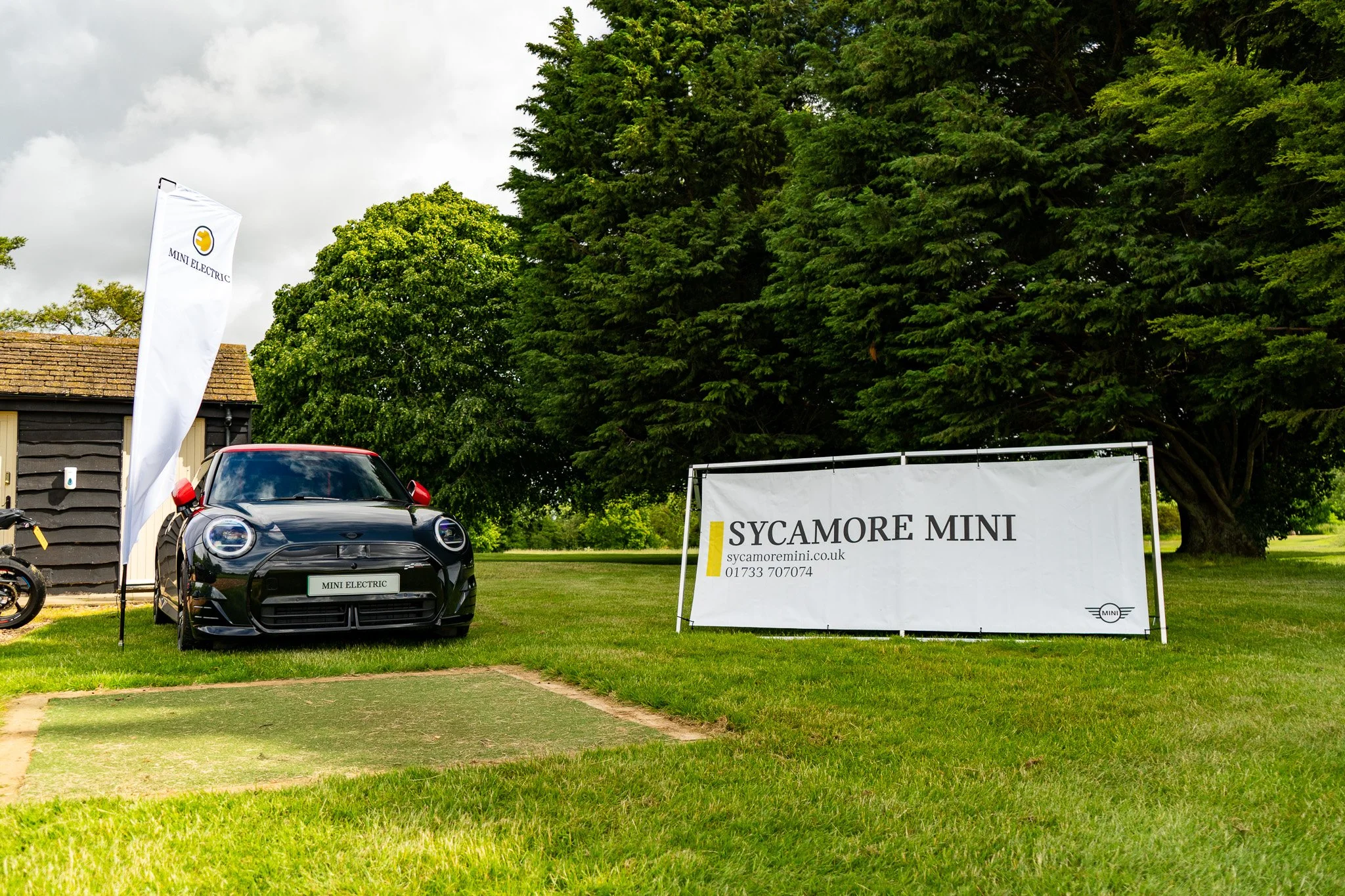 A black Mini Electric car parked on grass at a promotional event, with a white Mini Electric flag and a white banner displaying 'Sycamore Mini' and contact information, surrounded by green trees.