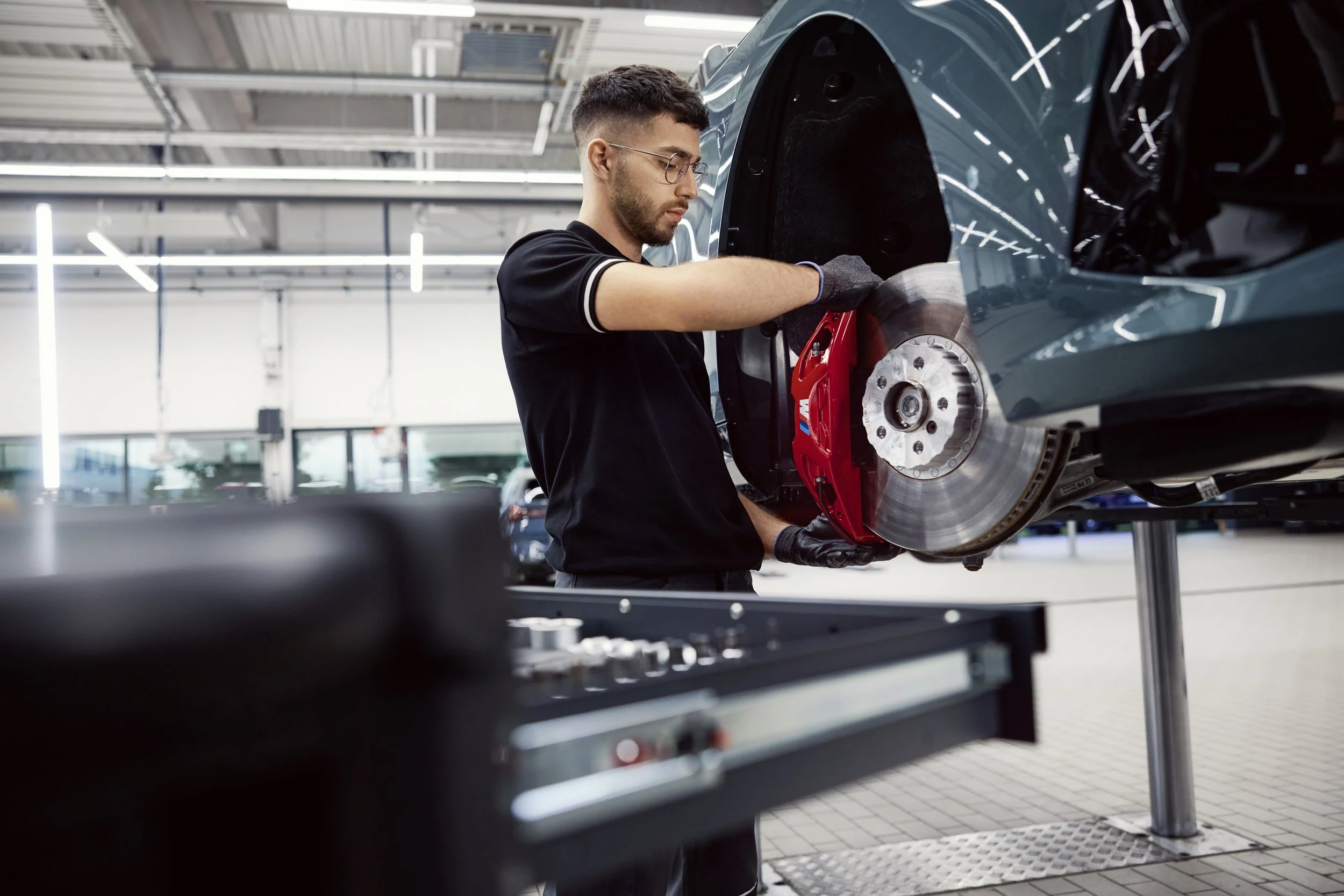 A man with glasses wearing gloves working on the brake system of a car that is lifted in an automotive repair shop.