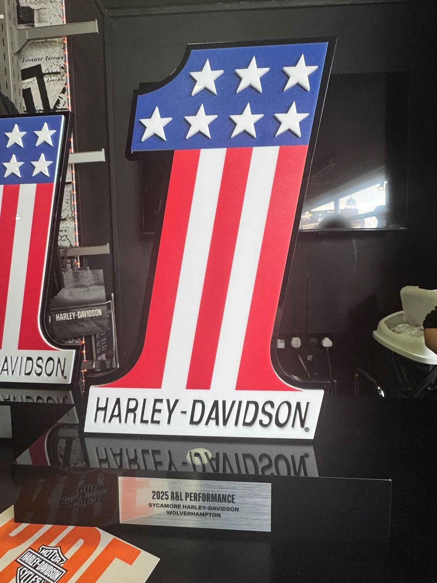 A large Harley-Davidson logo with an American flag theme, featuring white stars on a blue background with red and white stripes, displayed on a black surface. Smaller Harley-Davidson signs are also visible.