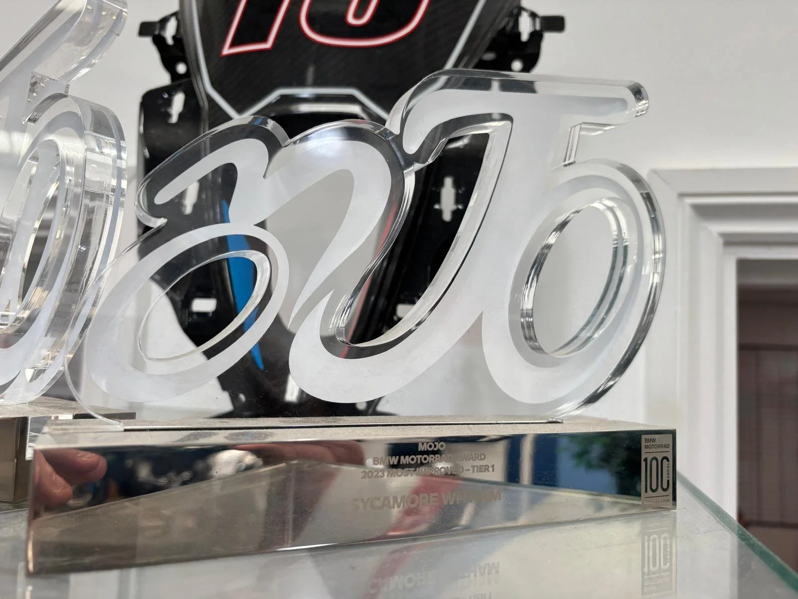 A clear acrylic 3D number '2023' displayed on a reflective metal base, with a blurred background showing a black and red sign and some electronic equipment.