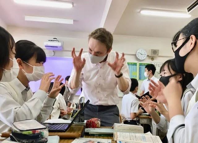 Teacher engaging students in classroom activity, all wearing masks, with textbooks and materials on desks.