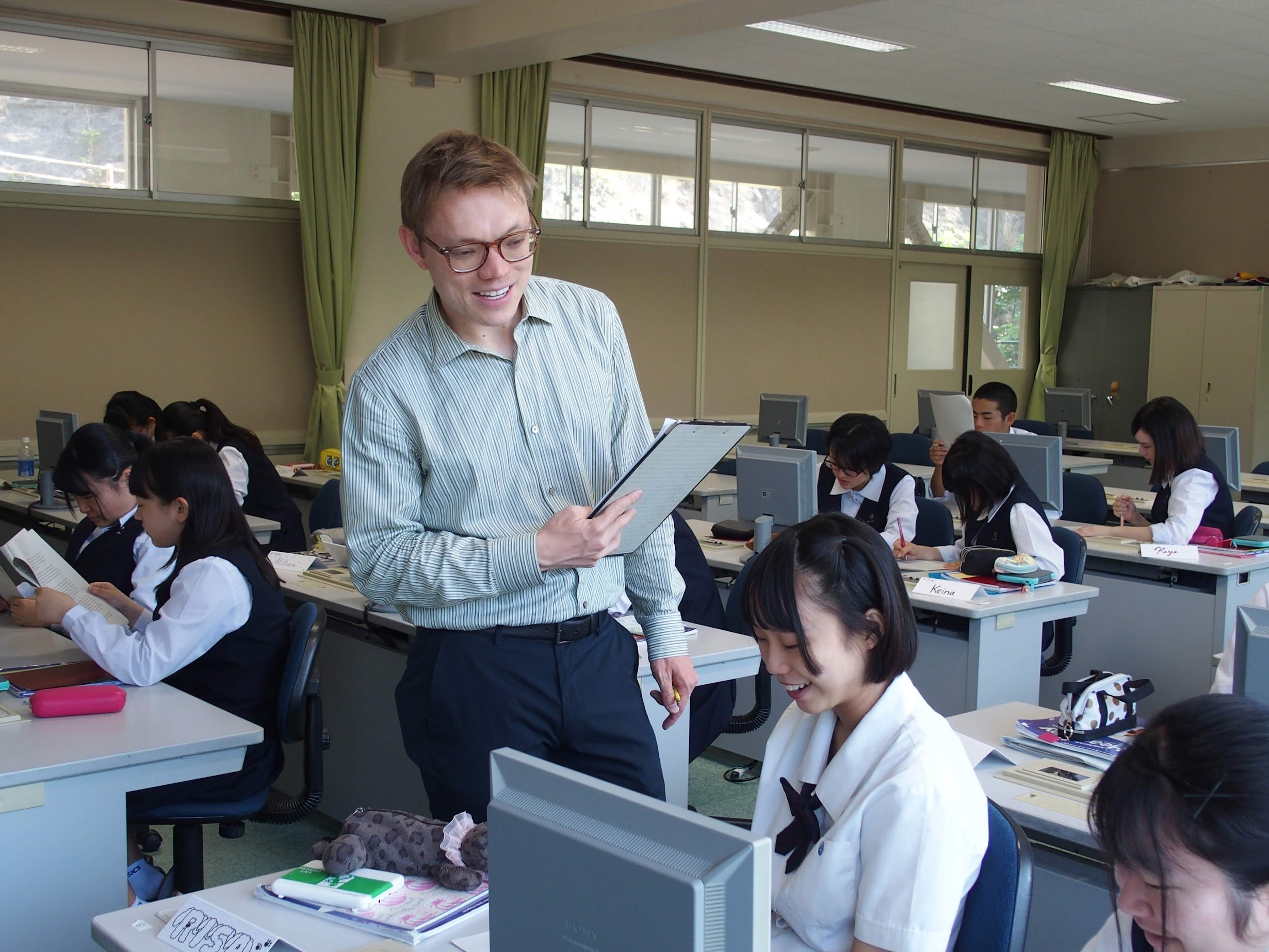 A classroom with students working at desks and a teacher smiling and looking at a tablet, surrounded by large windows with green curtains.