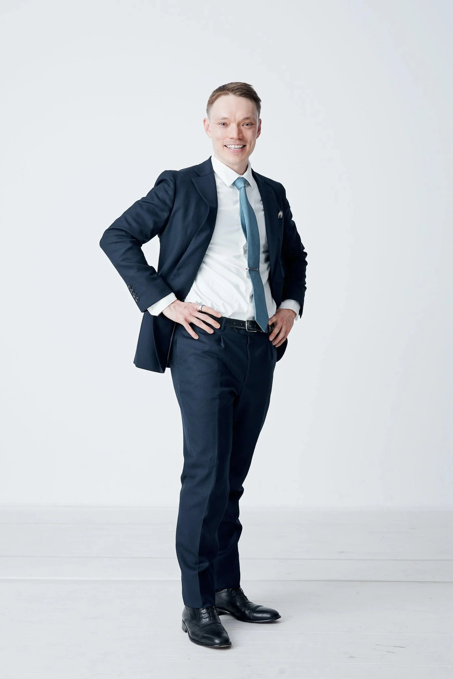 A young man in business attire standing confidently against a plain white background, with hands on hips and smiling.
