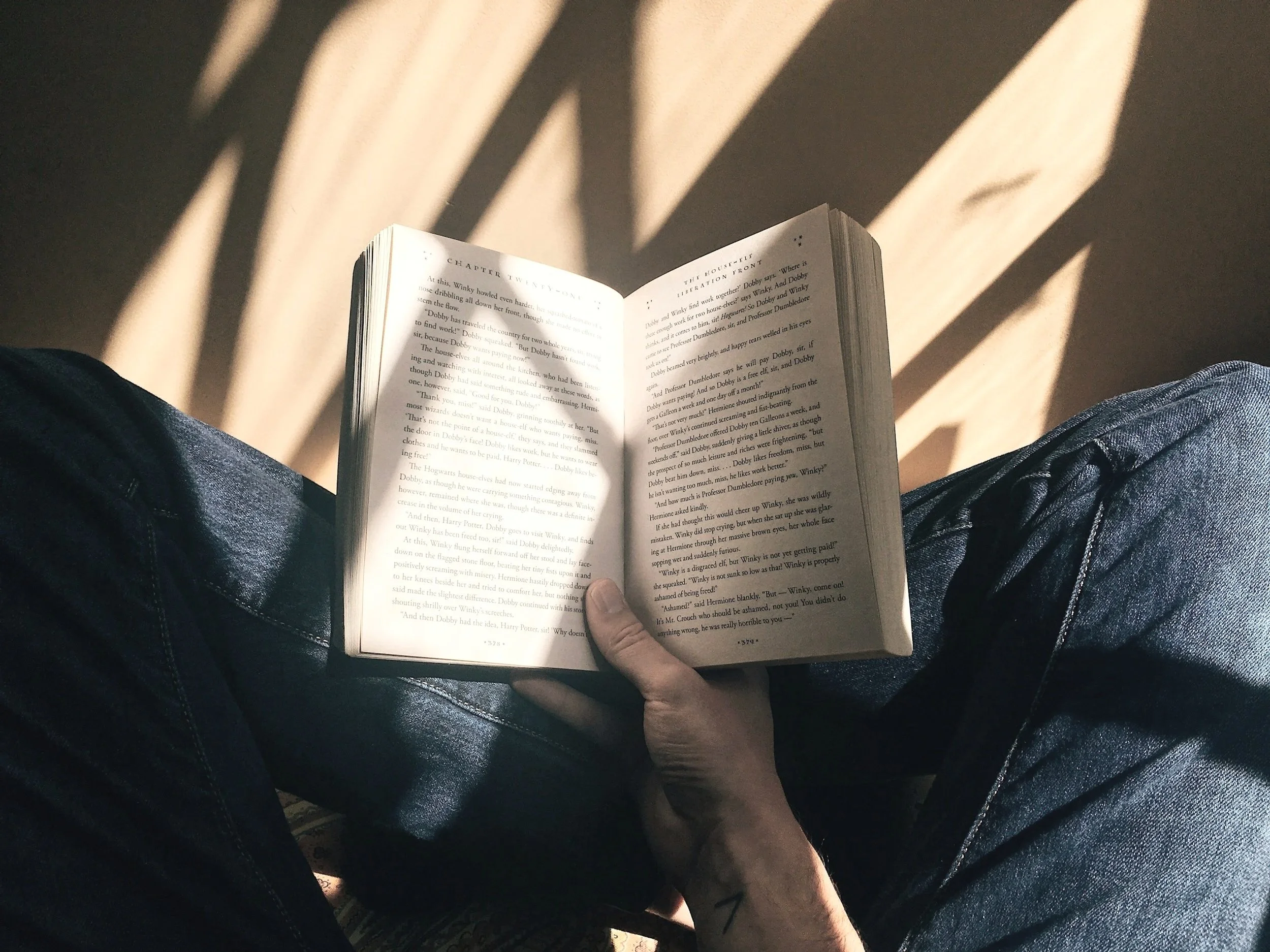 A person sitting with crossed legs wearing dark jeans, holding an open book, with sunlight creating shadows on the floor.