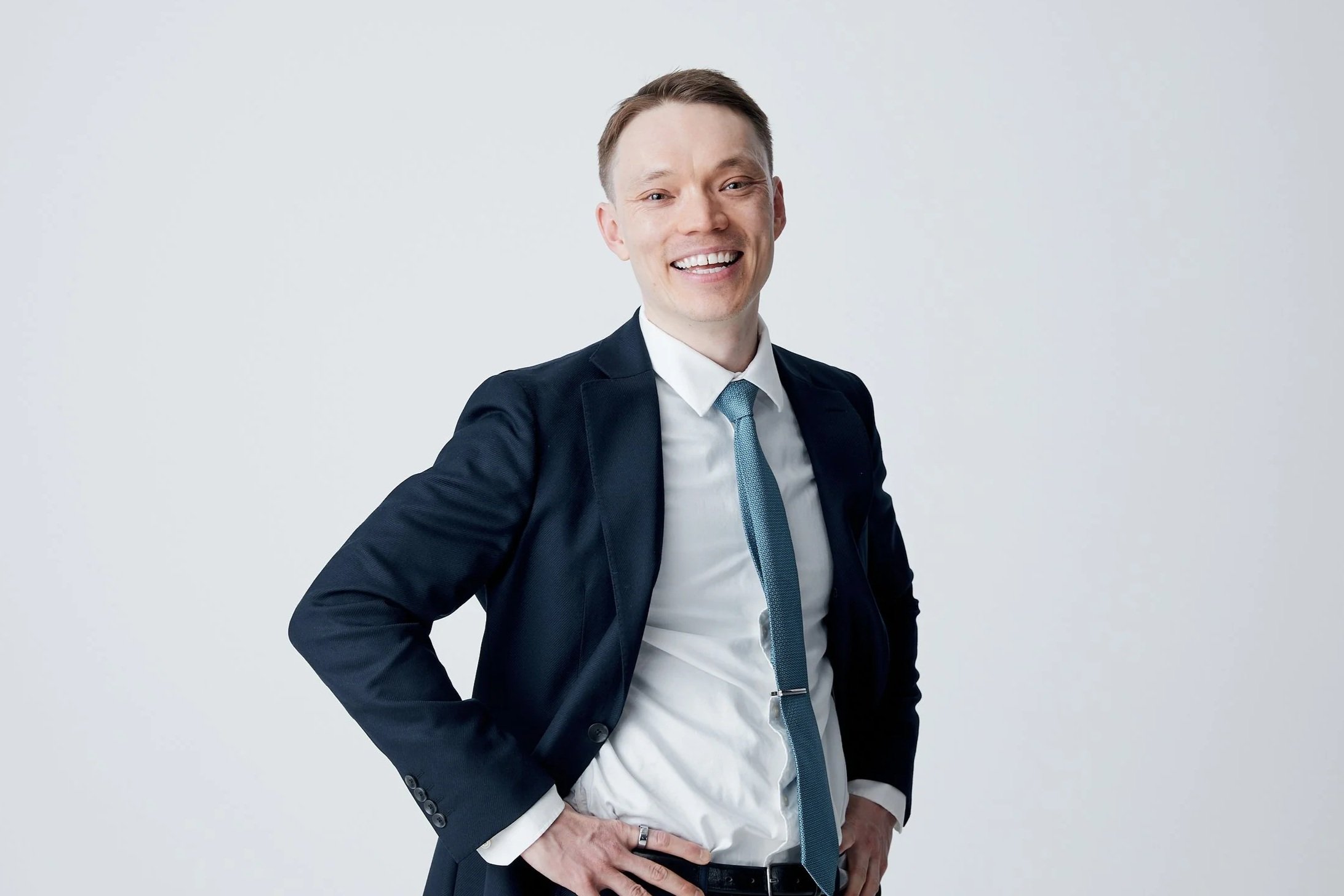 A smiling man in a dark suit with a light blue tie, standing against a plain light gray background.