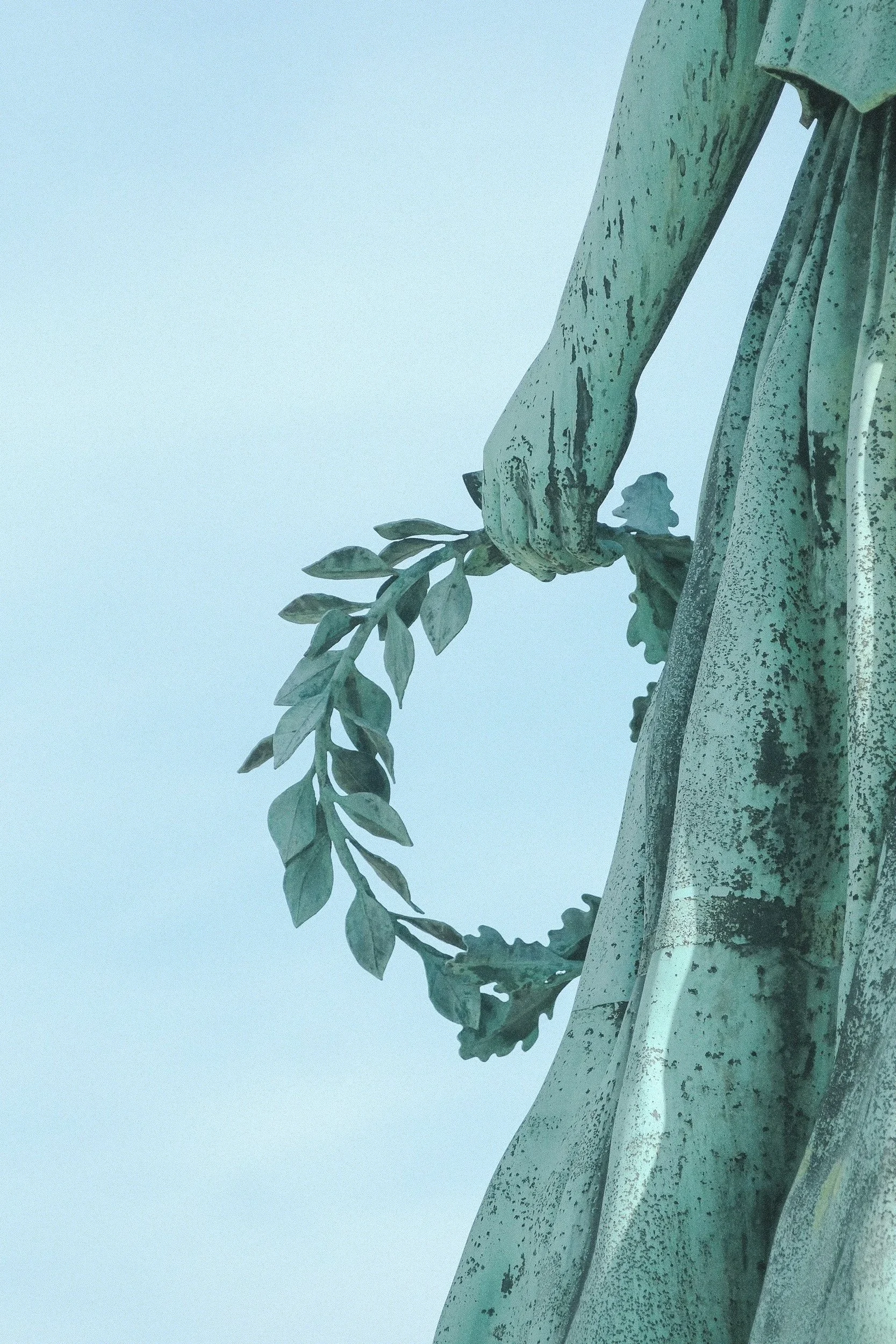 Close-up of the bottom part of a statue depicting a hand holding a branch with leaves. The statue appears to be made of metal with a weathered greenish patina. The background is a clear pale blue sky.