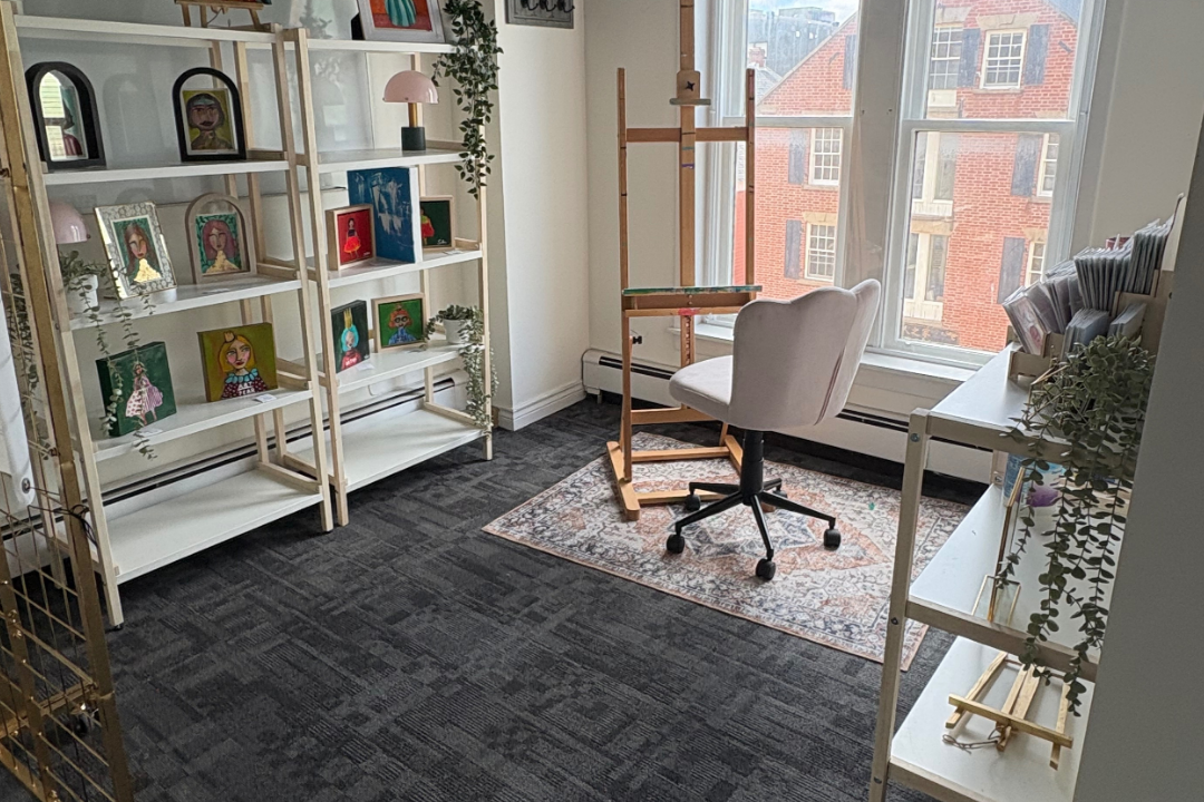 A cozy art studio with large windows, white bookshelves displaying colorful artworks, a white desk with art supplies, an adjustable wooden easel, and a white swivel chair on a patterned rug.