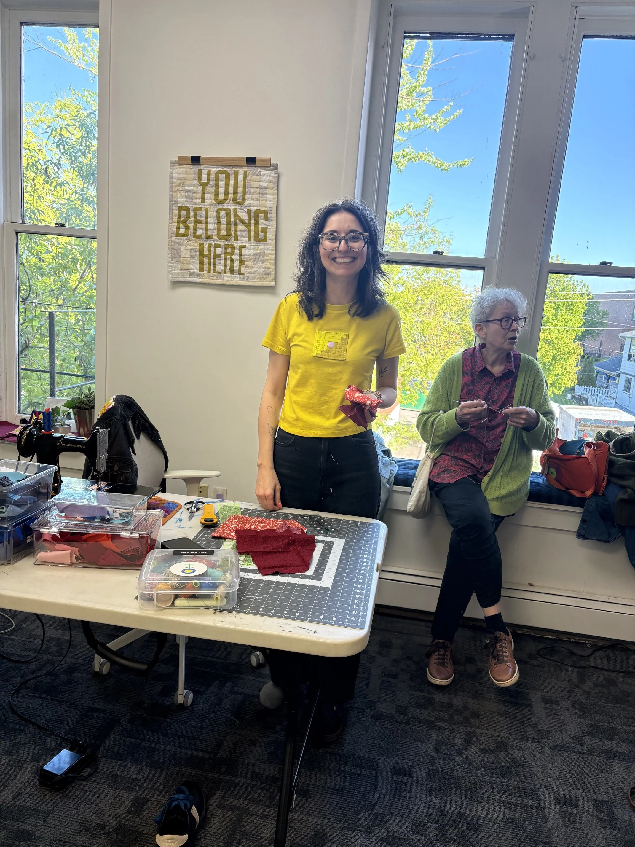 A woman with curly brown hair, glasses, and a yellow T-shirt smiling at the camera, holding a fabric piece in her left hand. Behind her, an older woman with curly white hair, glasses, and a green cardigan, is working with fabric. There are sewing supplies on a table, and a sign on the wall that reads "YOU BELONG HERE." Large windows in the background let in natural light and show trees and a building outside.