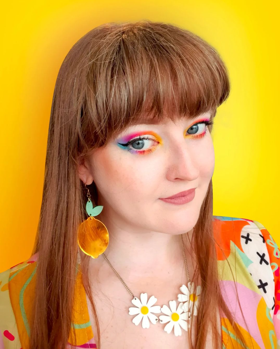 Portrait of Elyse Hendry wearing bright rainbow eye makeup, a colourful patterned top, a white daisy necklace and large lemon earrings against a yellow background.