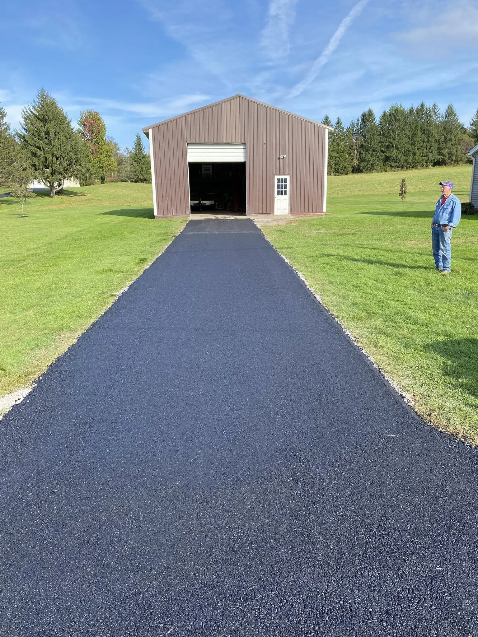A newly paved black asphalt driveway leading to a large brown barn with a white garage door and a white side door. A man in blue jeans, a blue jacket, and a baseball cap stands on the right side of the driveway. The sky is blue with some wispy clouds, and the surrounding area is grassy with trees.