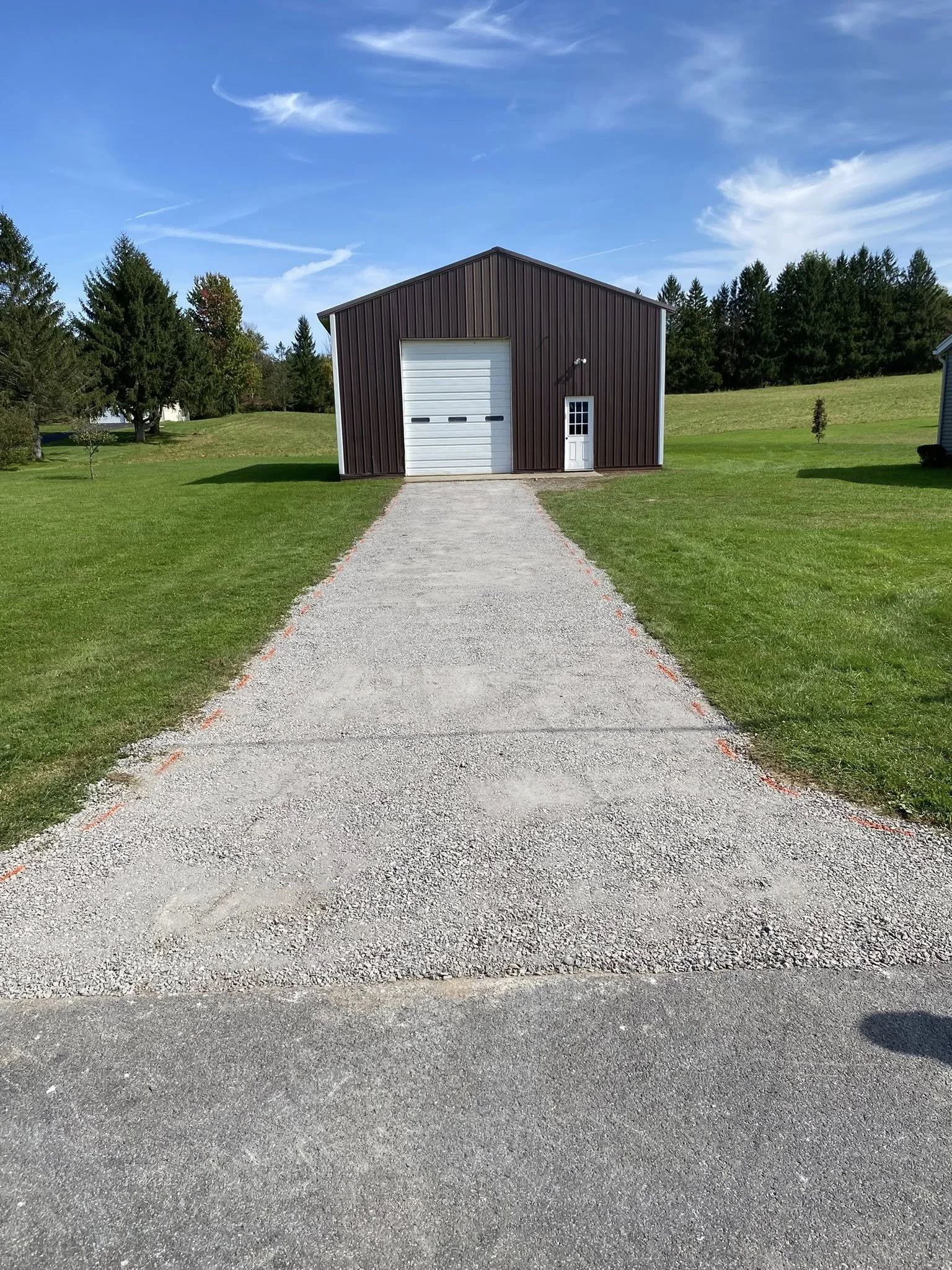 A gravel driveway leading to a metal-sided shed with a white garage door and a small white door, set in a grassy, open area with trees and a blue sky.