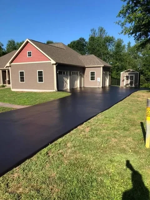 Newly paved black asphalt driveway leading to a double garage of a house with beige and pink siding and a small shed in the backyard.
