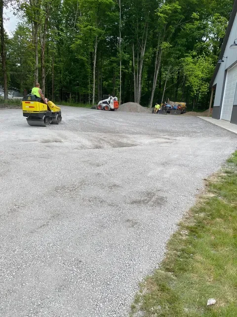 A construction site with a worker operating a small road roller, a pile of gravel, and a small excavator or loader, surrounded by a wooded area and a building with a garage door.