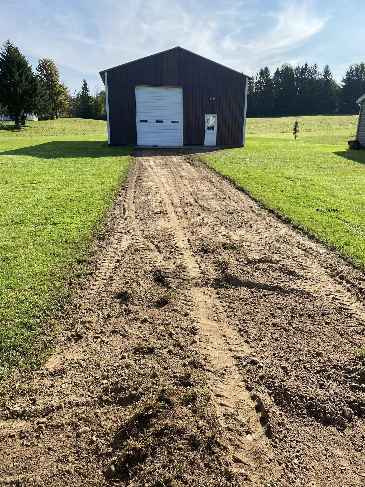 A dirt driveway leading to a large metal shed with a white garage door and a smaller white door, surrounded by green grass and trees in the background under a partly cloudy sky.