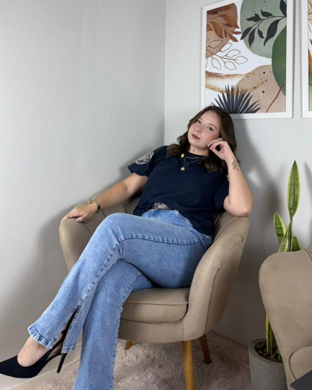 Mulher sentada em cadeira confortável, com cabelos castanhos, vestindo camisa azul, jeans e sapatos pretos, em uma sala decorada com quadros e plantas.