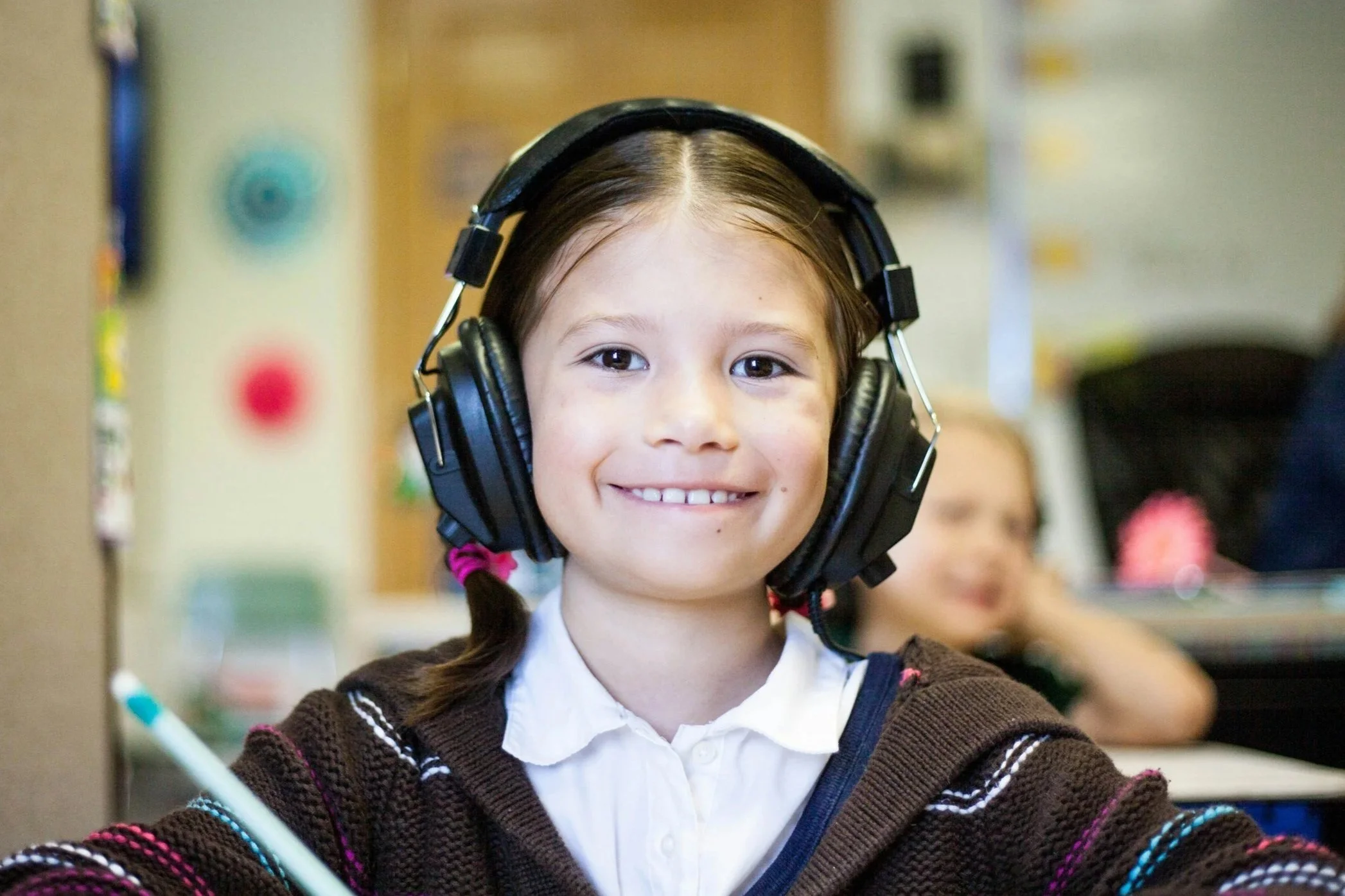 A young girl with brown hair in pigtails, wearing a brown sweater and white shirt, smiling while wearing large black headphones in a classroom.