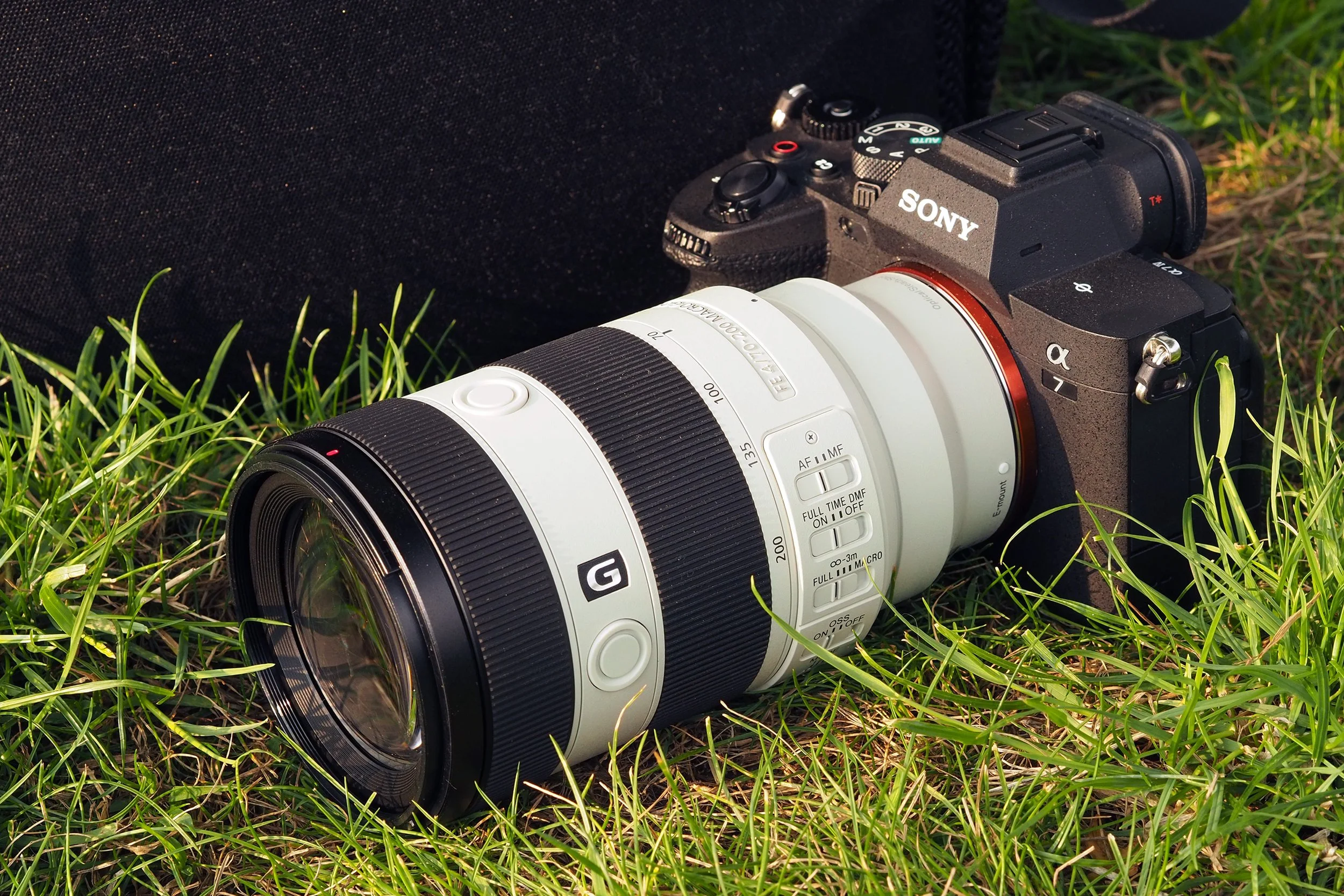 A Sony mirrorless camera with a white zoom lens lying on green grass, with a black carrying case in the background.
