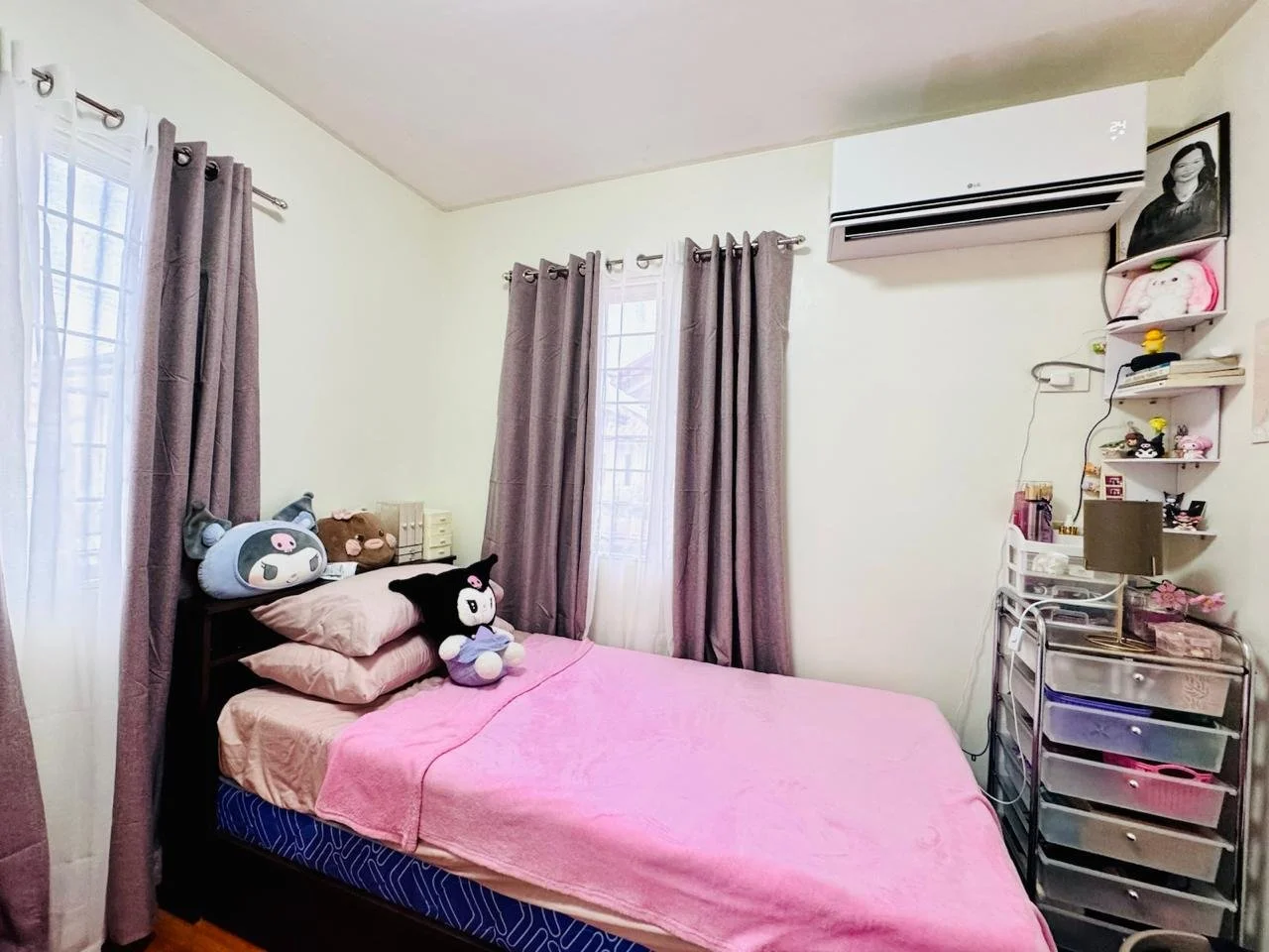 Bedroom with a bed covered in pink blanket, plush toys on pillows, purple curtains, white wall air conditioner, shelf with plush toys and decorative items, gray storage cart with drawers, and windows with white and purple curtains.