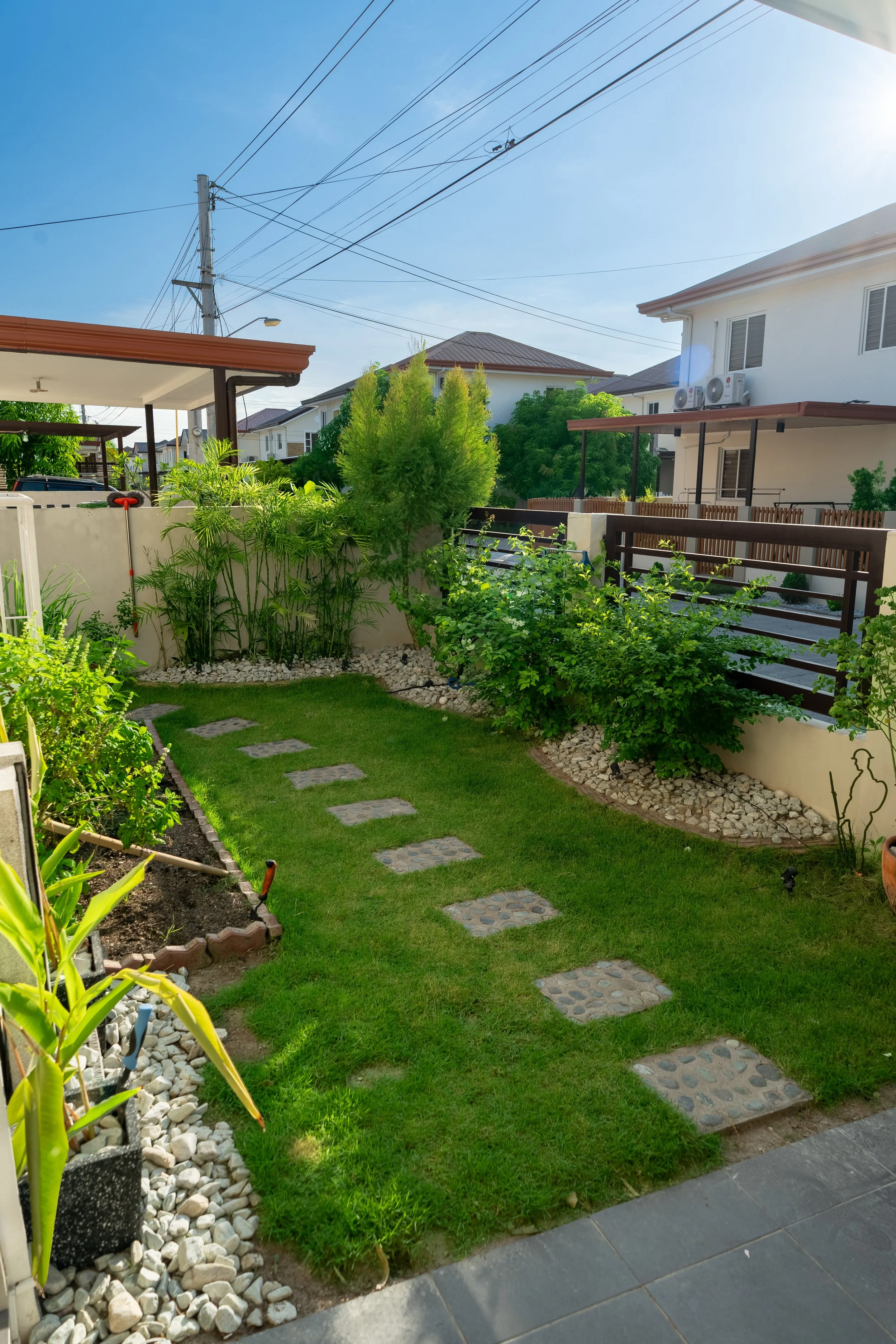 A well-maintained residential backyard with a stone pathway, lush green lawn, various plants, and trees, surrounded by a white wall and neighboring houses. Overhead power lines are visible in the bright, sunny sky.
