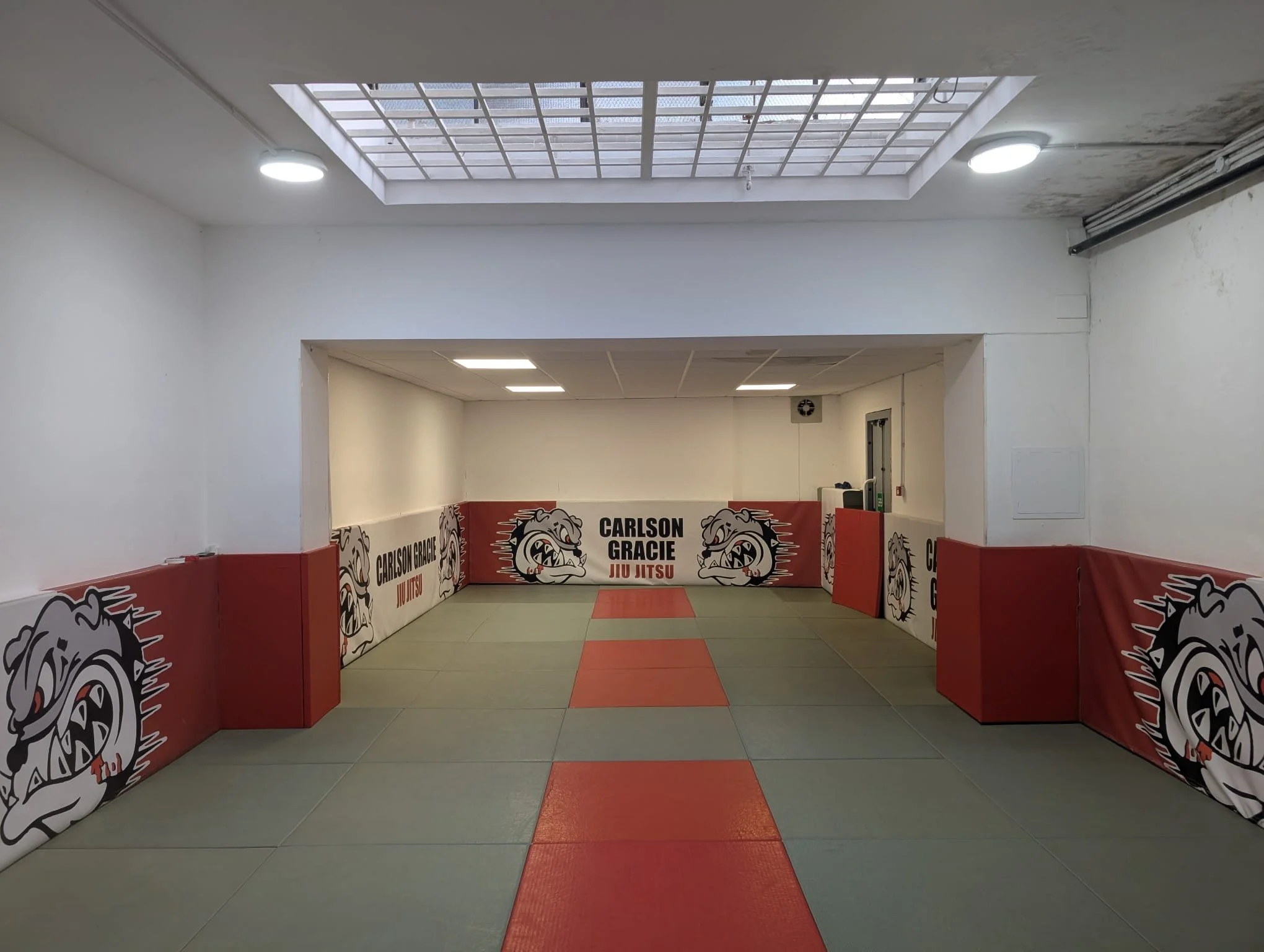 Training dojo with a padded red and gray mat, surrounded by padded barricades featuring bulldog logos and text for Carlson Gracie Jiu Jitsu, under a ceiling with skylight and bright lights.